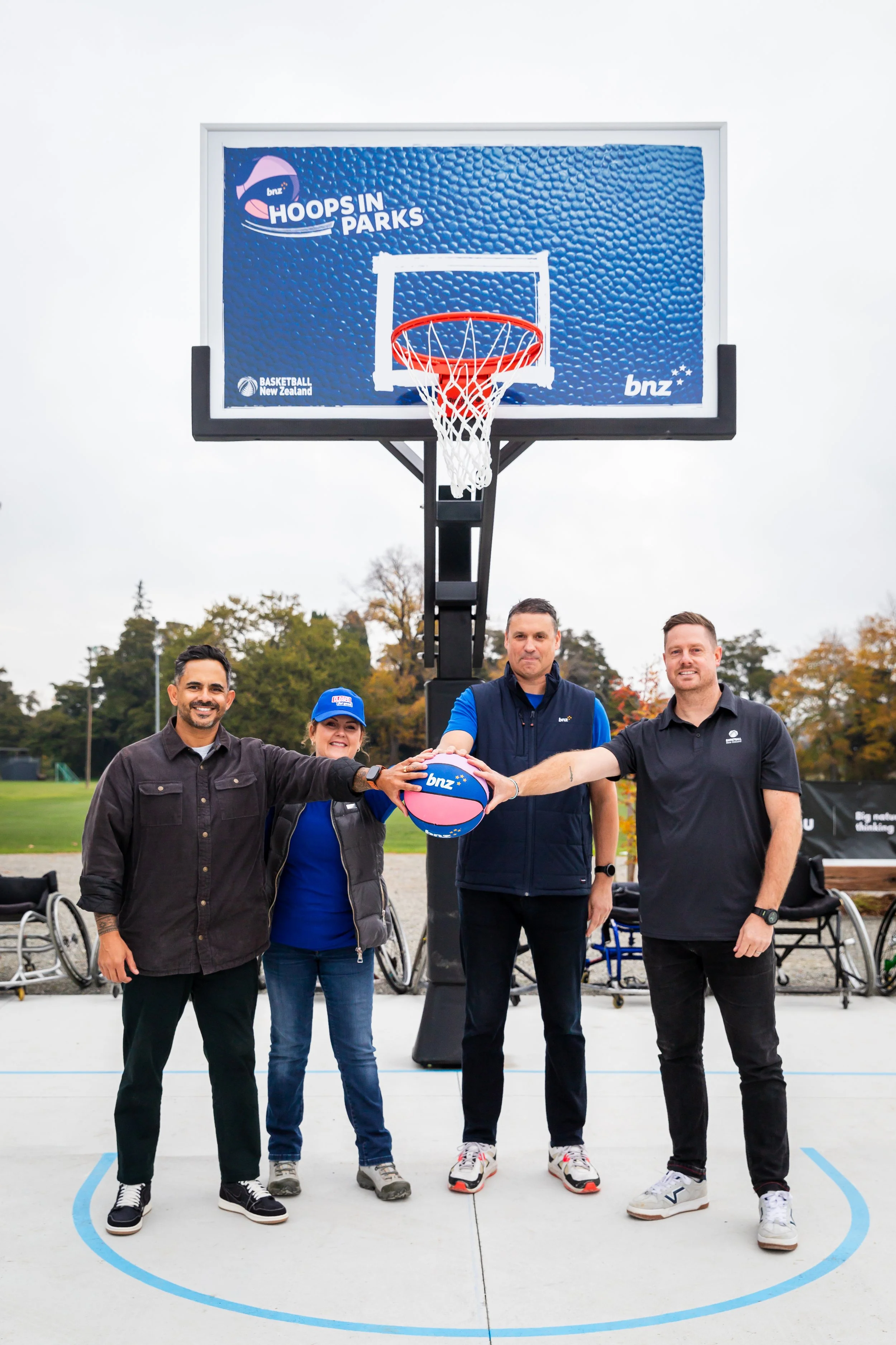 Group of five diverse people standing on a basketball court outside, participating in a basketball event with a basketball and a hoop. They are smiling and holding hands together, with a banner in the background that says "BNZ Hoops in Parks." The ev