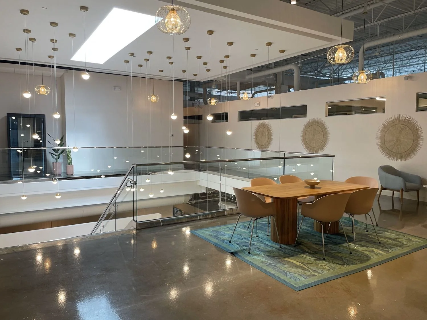 Interior of a modern, well-lit office or meeting space with a wooden table, beige chairs, and a patterned rug. A glass balcony with metal railing overlooks a lower level. Decorative wall art and hanging pendant lights add to the ambiance.