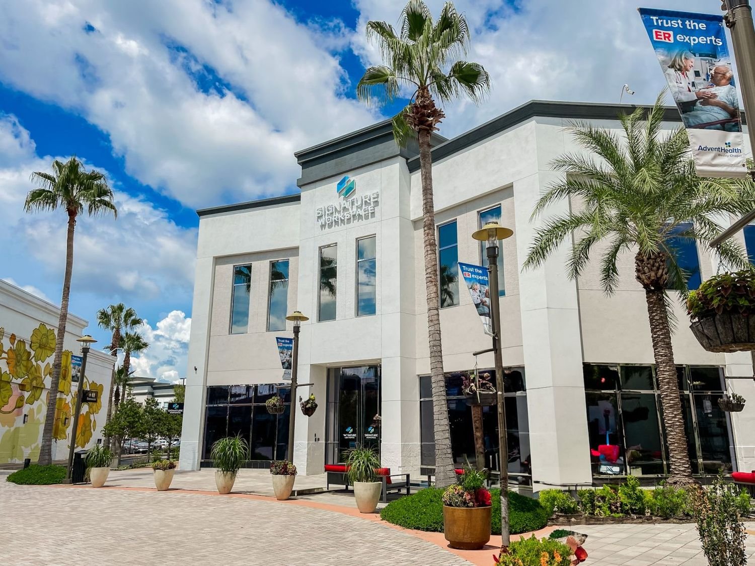 Exterior view of a modern white building named 'Signature Workspace' with tall windows, palm trees, street lamps, potted plants, and banners that read 'Trust the ER experts,' under a partly cloudy blue sky.