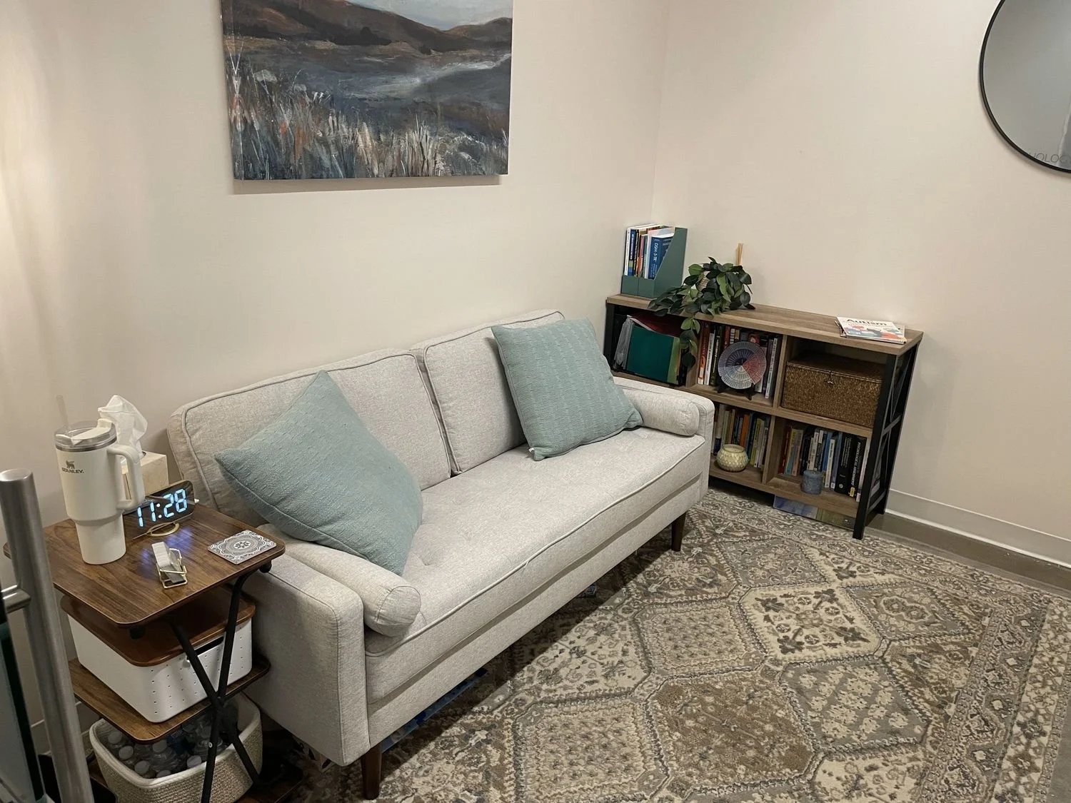 A cozy living room corner featuring a light gray sofa with two matching blue pillows, a wooden side table with a clock displaying 1:28, a white diffuser, and a tissue box. Behind the sofa is a framed abstract landscape painting, and to the right is a wooden bookshelf with various books, a potted plant, and decorative items. The floor is covered with a patterned area rug.