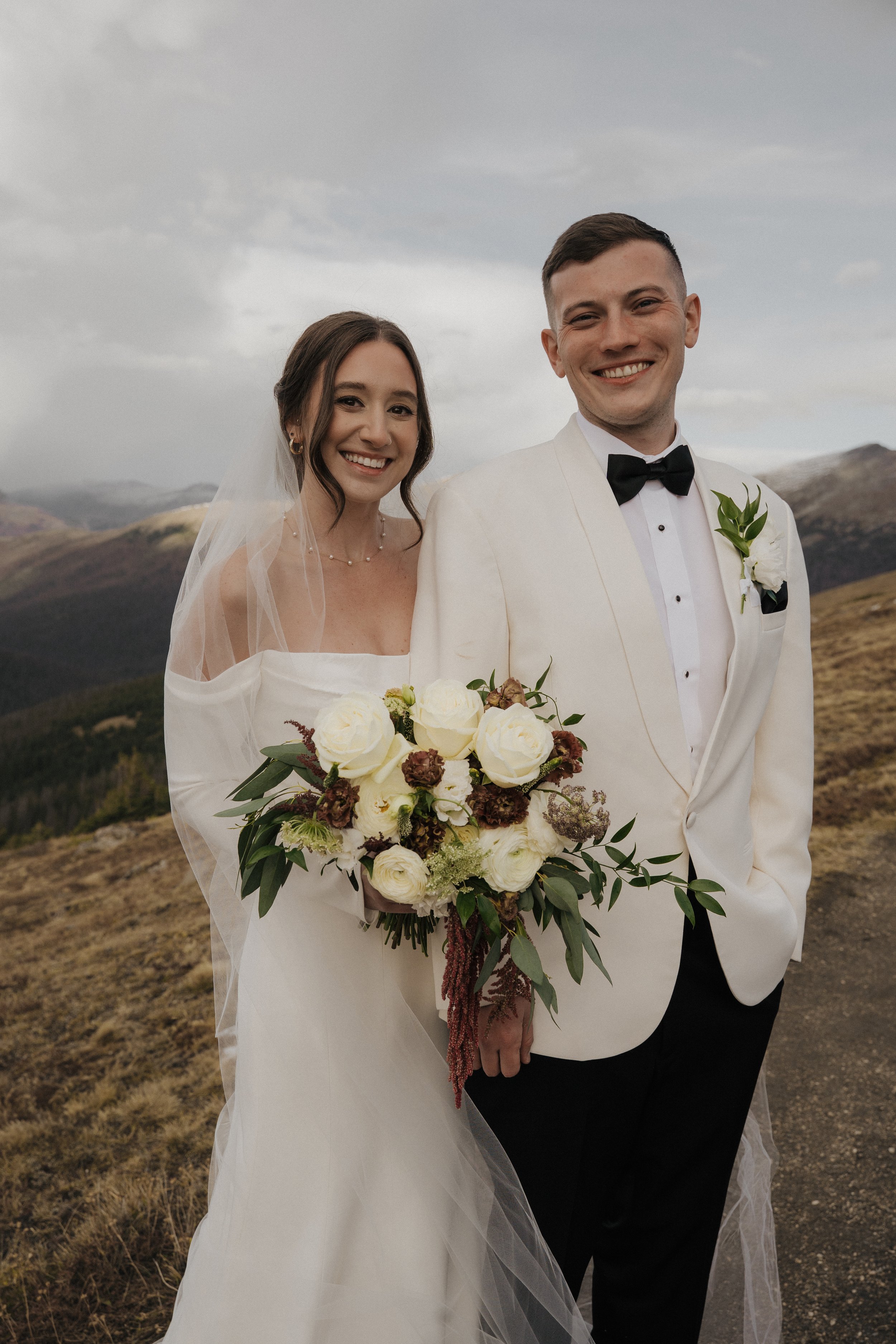 A bride and groom smiling outdoors during their wedding, with mountains and cloudy sky in the background. The bride is holding a bouquet of white and dark red flowers, and the groom is dressed in a white tuxedo with a black bow tie.