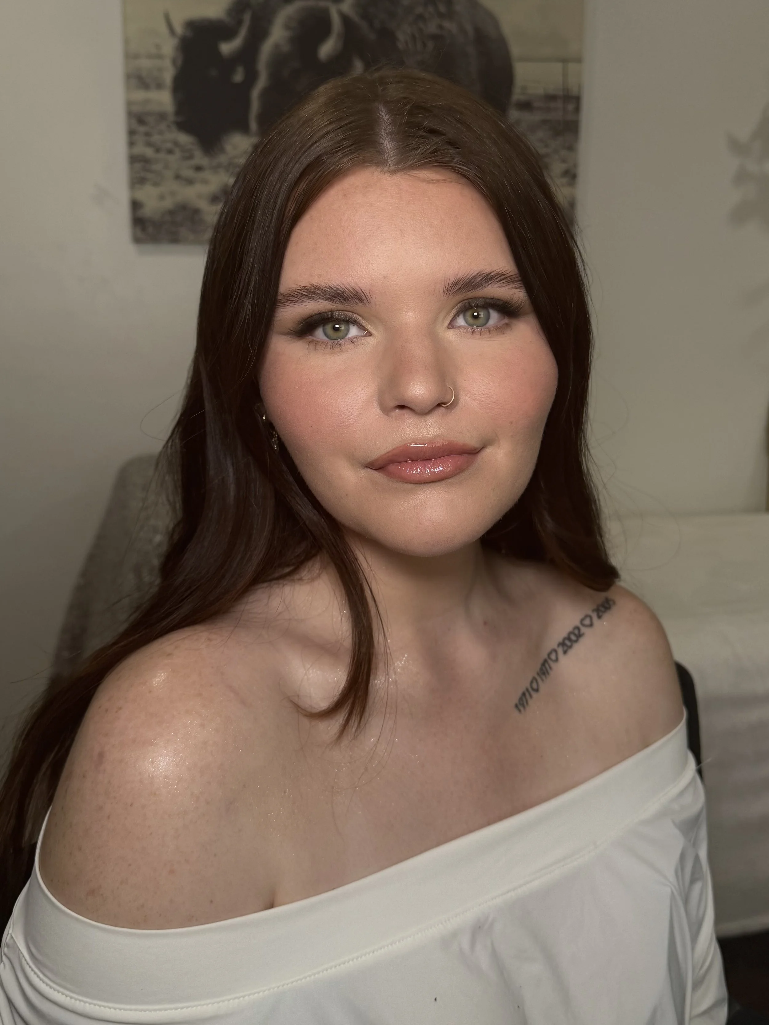 Close-up of a woman with long brown hair, green eyes, and a nose ring, smiling slightly and wearing an off-shoulder white top, with a black-and-white animal print poster on the wall behind her.