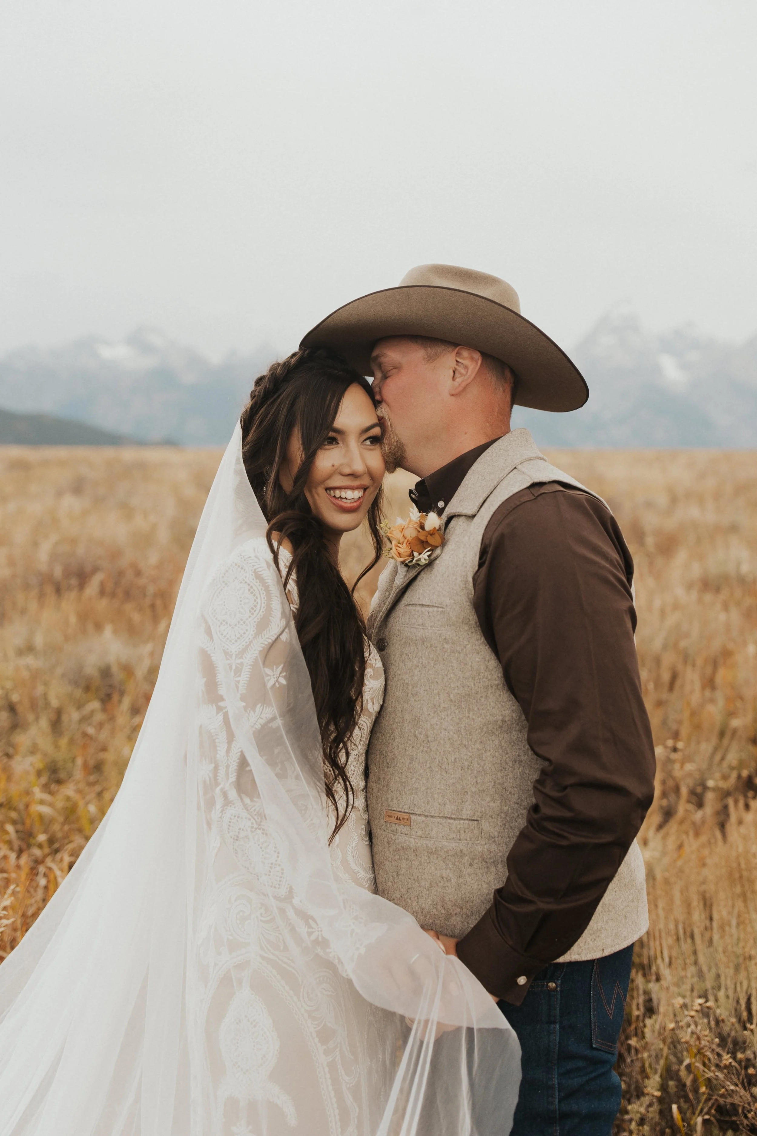 A bride and groom in a field, with the groom kissing the bride on the forehead.