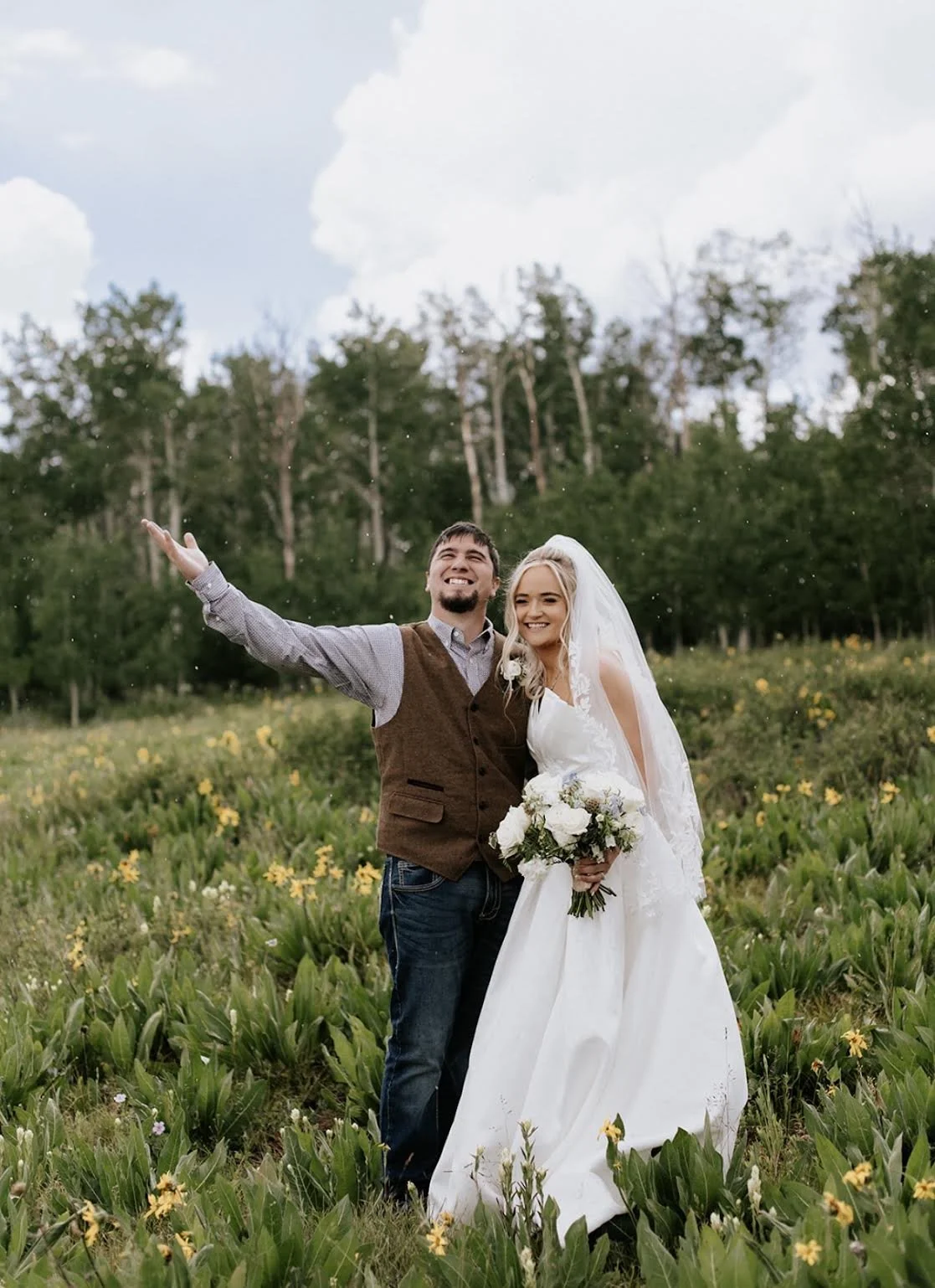 A newlywed couple standing in a field of yellow flowers, smiling happily, with the groom raising one arm and the bride holding a bouquet of white flowers, outdoors with trees and a cloudy sky in the background.