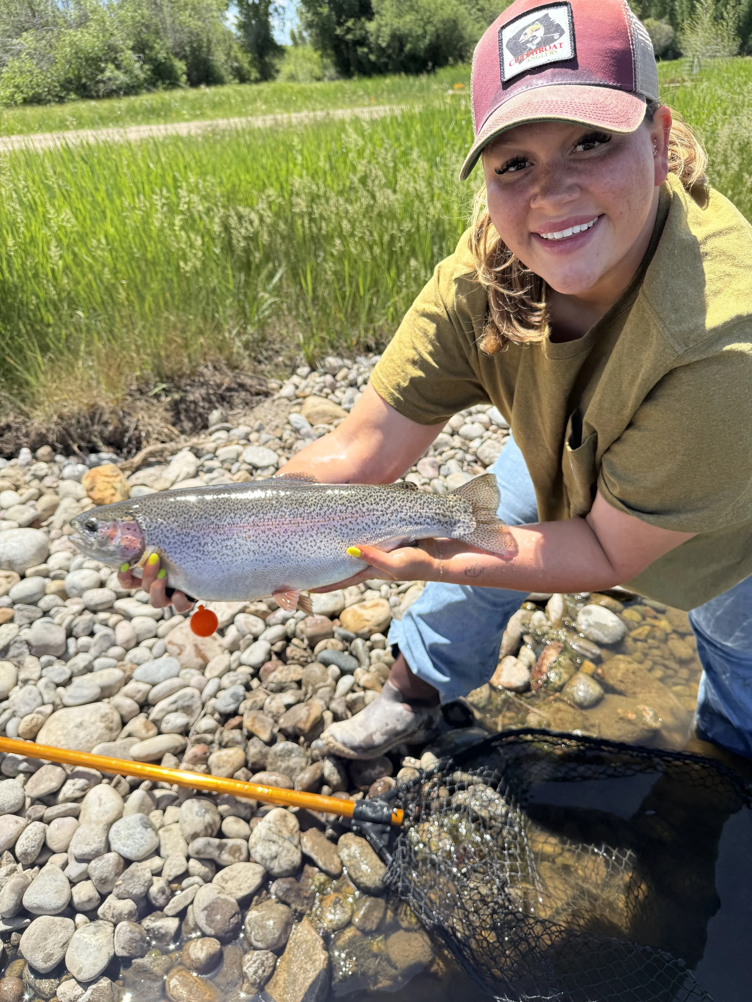 A woman with a red and beige cap and a yellow shirt is holding a rainbow trout near a rocky riverbank with green grass and trees in the background.