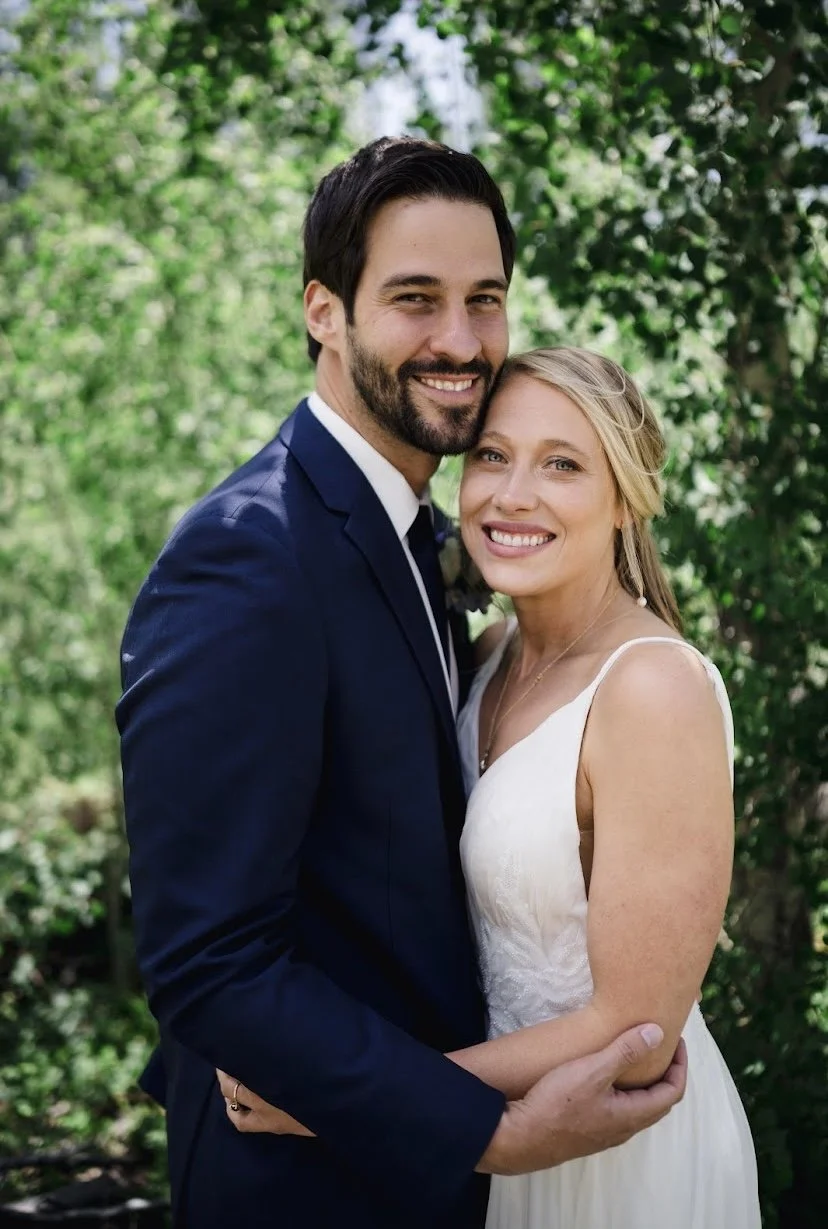 A happy couple dressed in wedding attire, standing outdoors with lush greenery in the background, smiling and embracing.