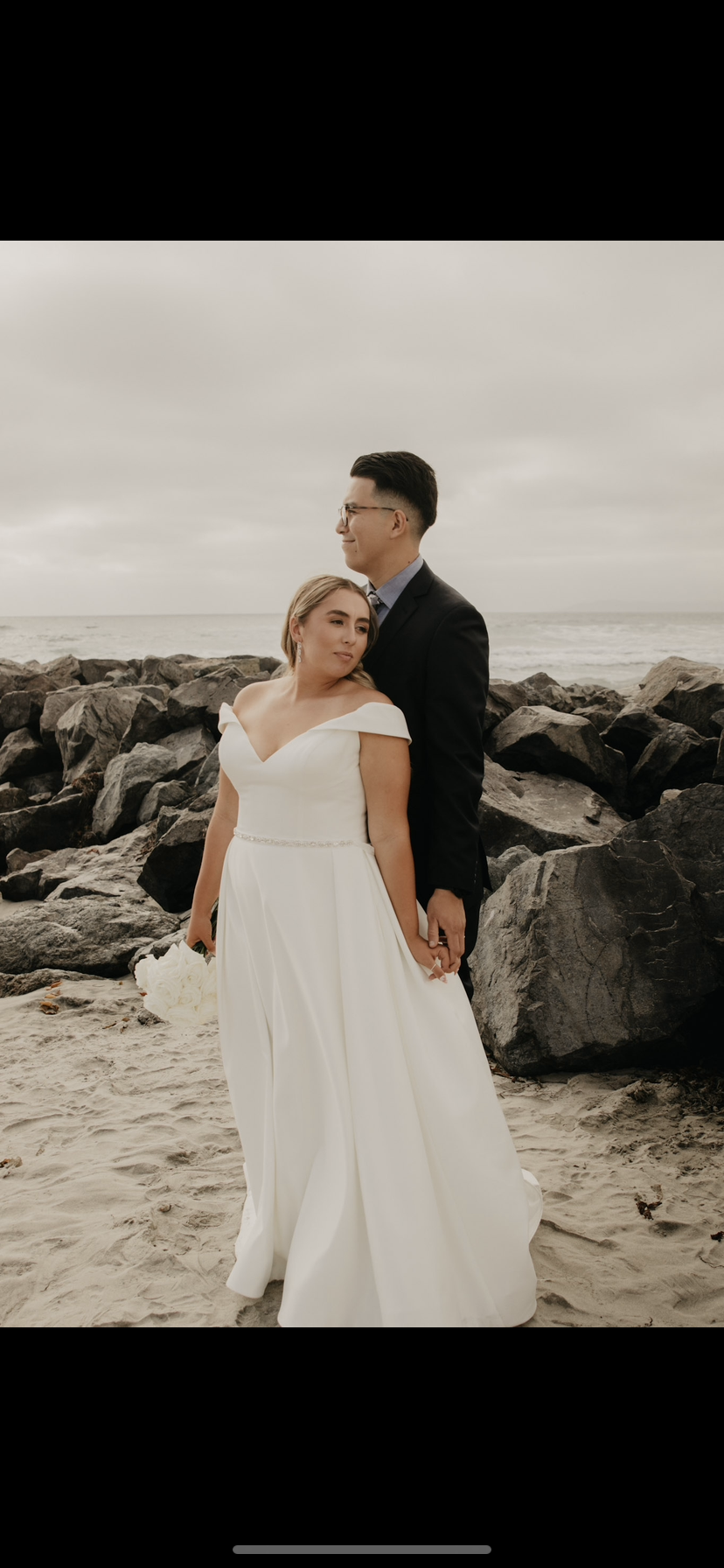 A bride and groom on a rocky beach, dressed in wedding attire, holding hands, with an overcast sky and the ocean in the background.