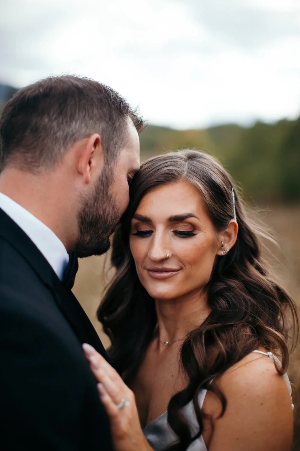 A man and woman sharing an intimate moment outdoors, with the man leaning his forehead against the woman's forehead, both with closed eyes and gentle expressions.