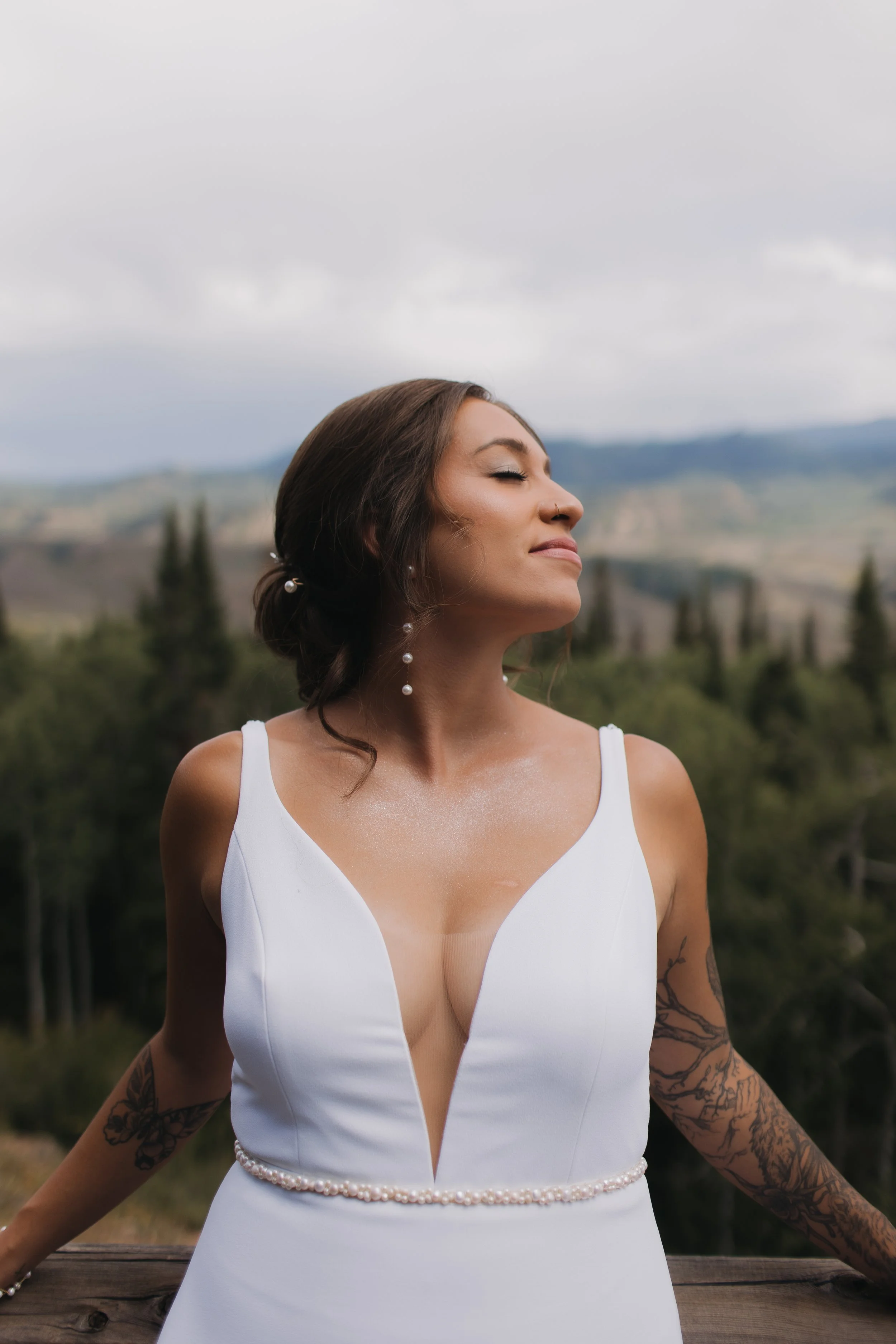 A woman in a white dress with pearl details stands outdoors with her eyes closed and a peaceful expression, surrounded by nature and a cloudy sky.