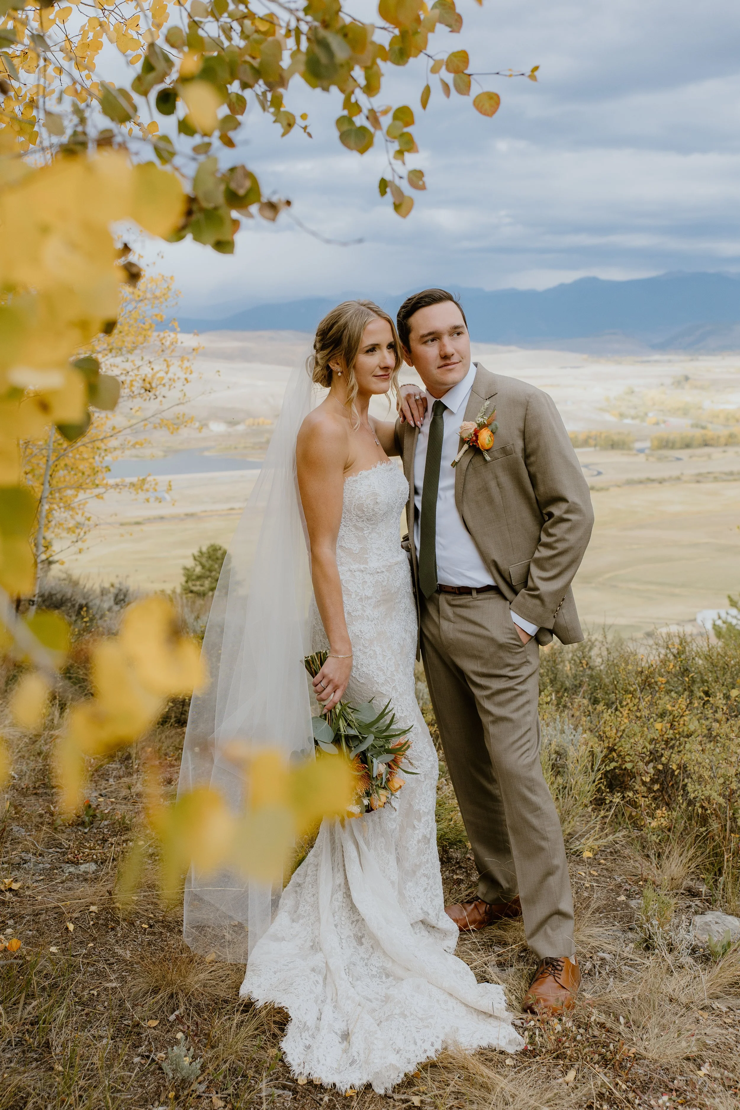 A bride and groom pose outdoors on a fall day with yellow leaves in the foreground and mountains in the background, for a wedding photo.
