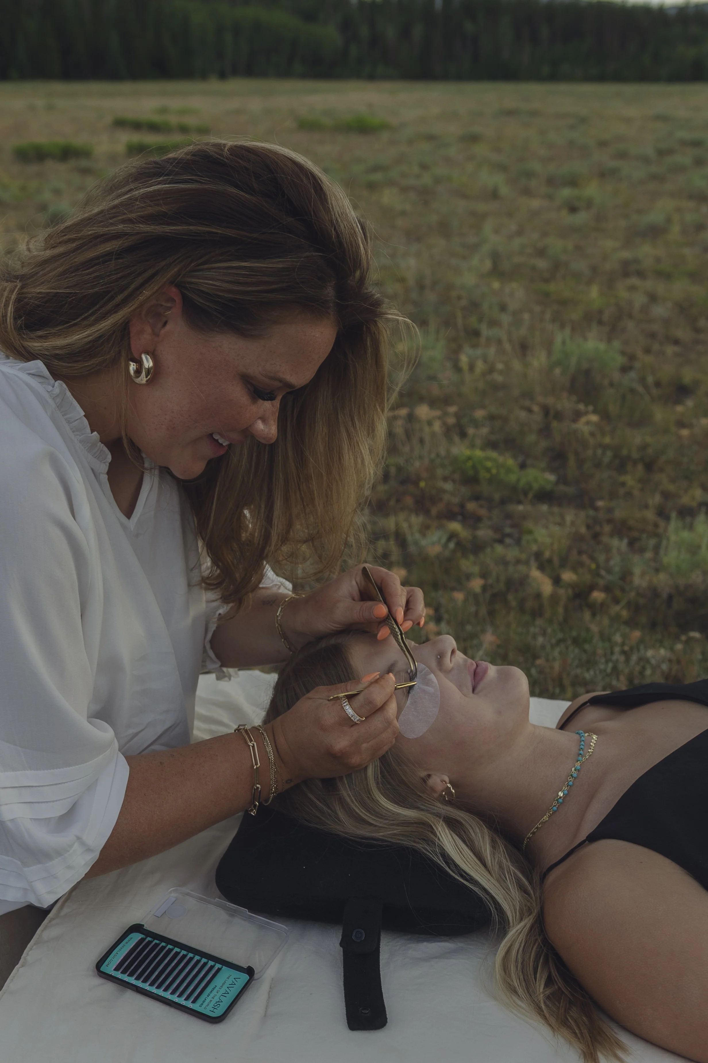 A woman is lying on a table with her eyes closed while another woman, standing, applies makeup or skincare to her face outdoors. The background shows grassy land and trees.