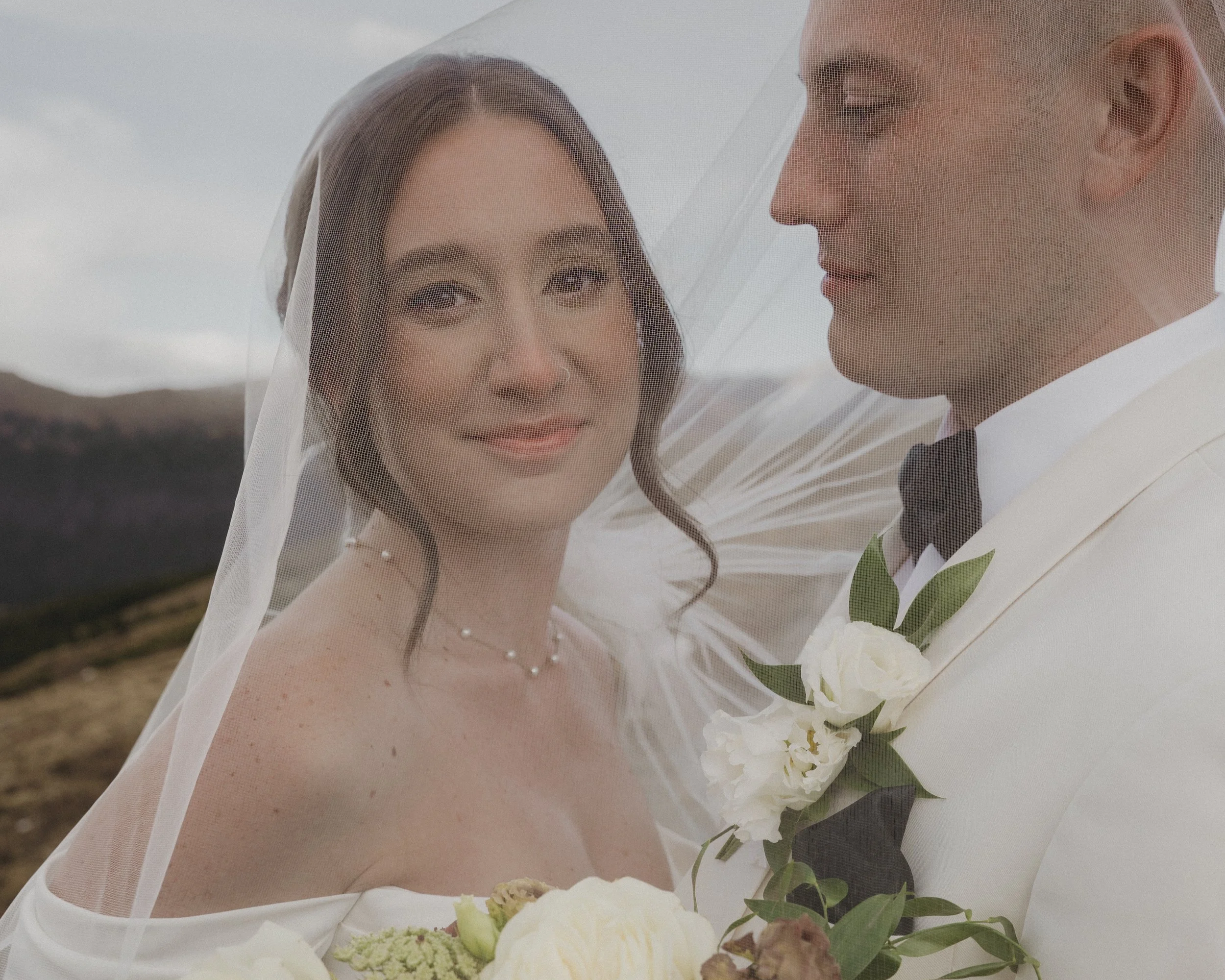 A bride and groom looking at each other under a wedding veil outdoors with mountains and cloudy sky in the background.