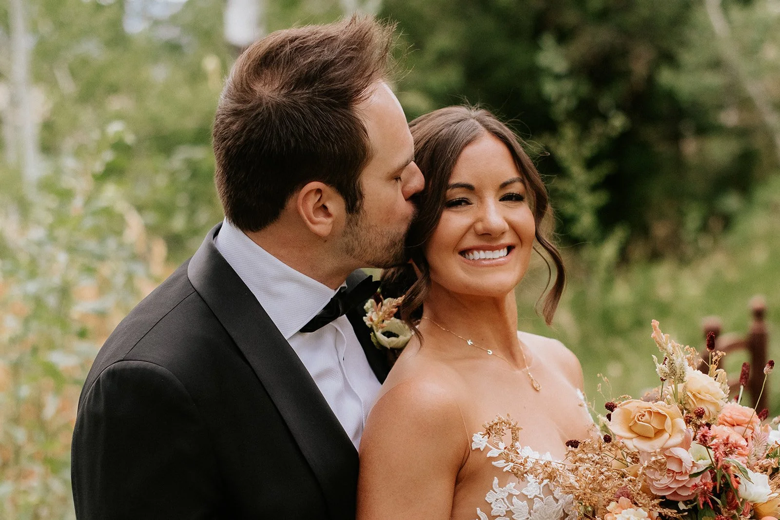 A groom in a tuxedo kisses a smiling bride in a strapless wedding gown holding a bouquet of flowers, outdoors with greenery in the background.