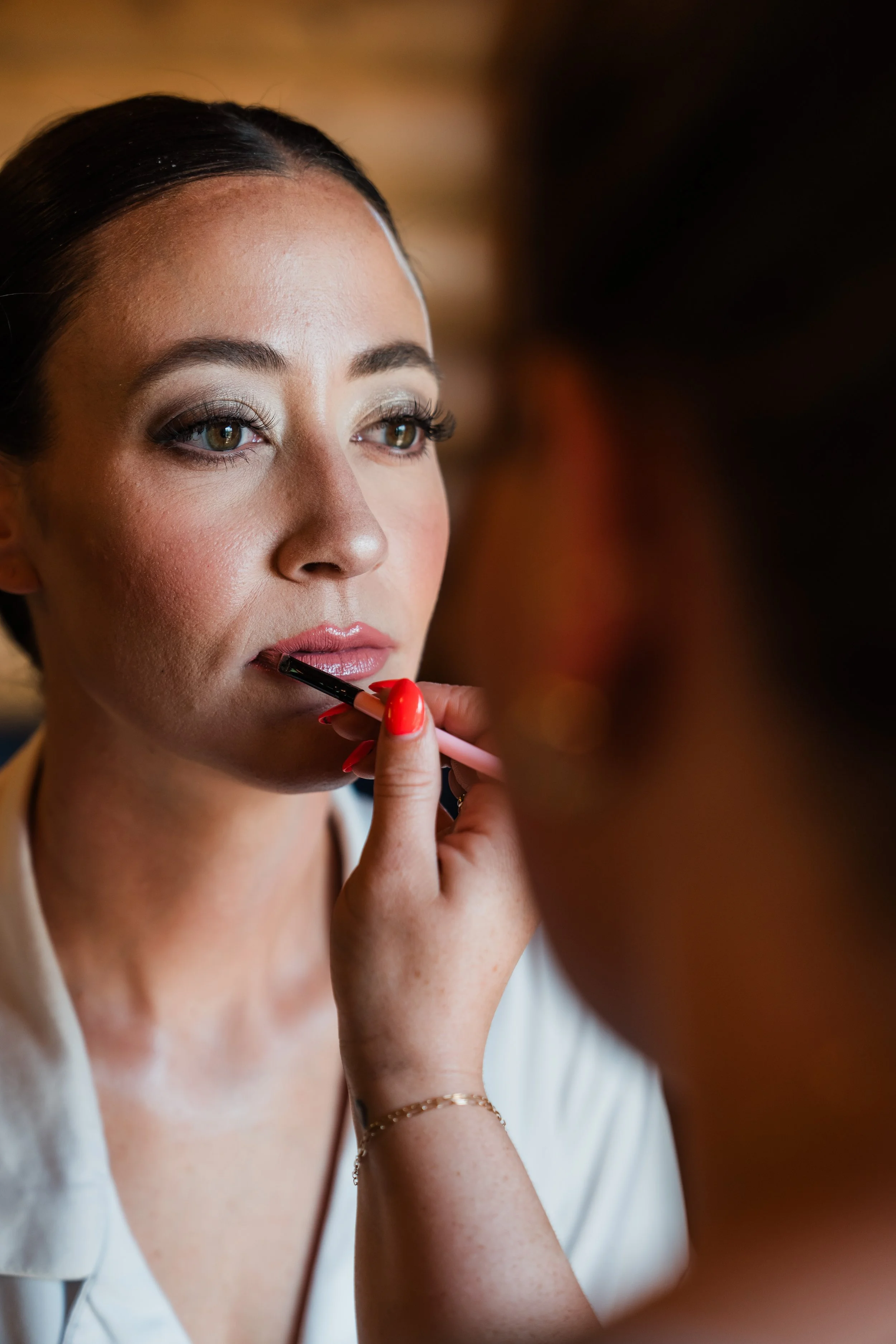 A woman with fair skin and makeup, applying lipstick with a brush, wearing a white top, and gold jewelry on her wrist.