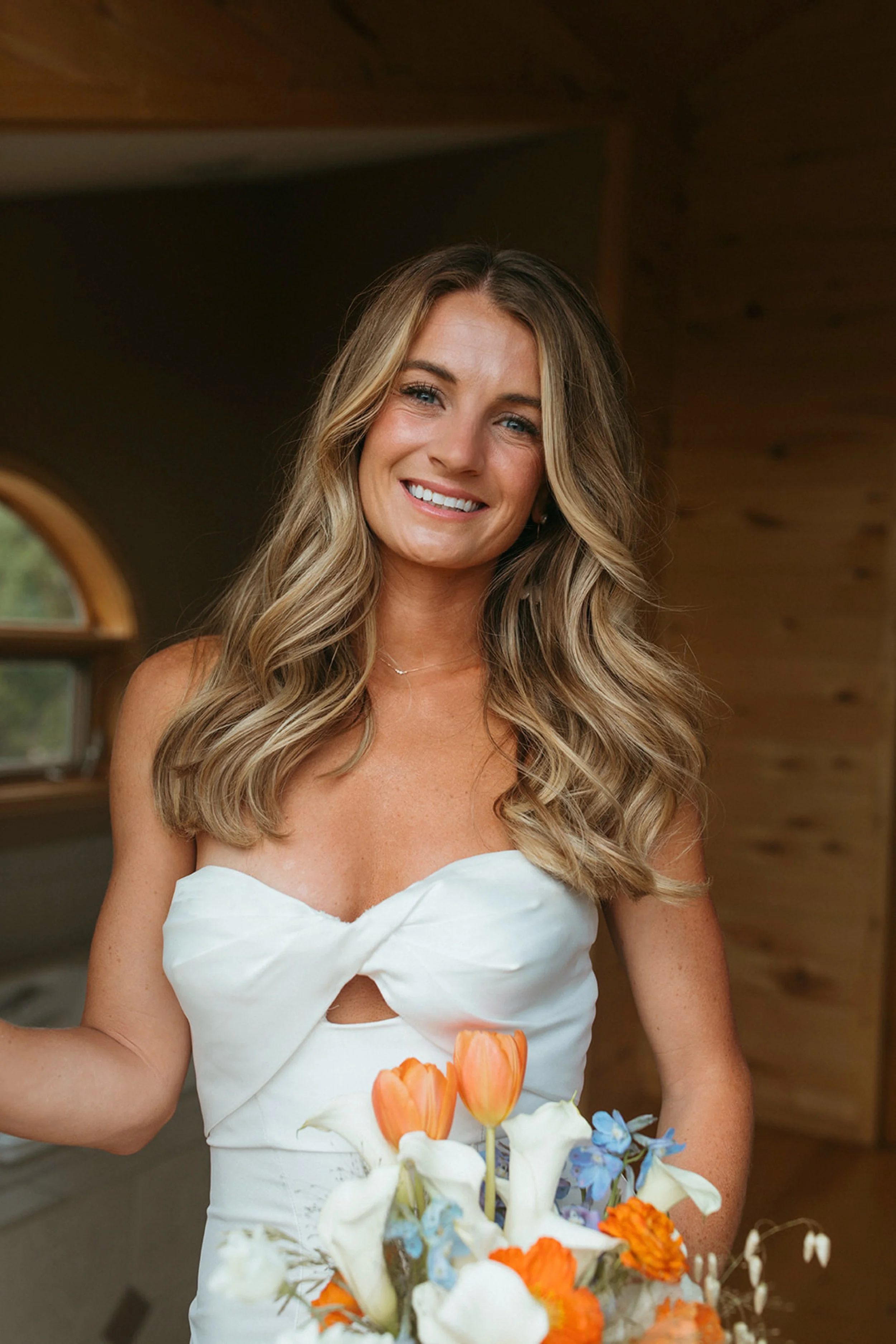 A smiling woman with long wavy blonde hair wearing a white strapless dress, holding a bouquet of white, orange, and blue flowers inside a wooden room.