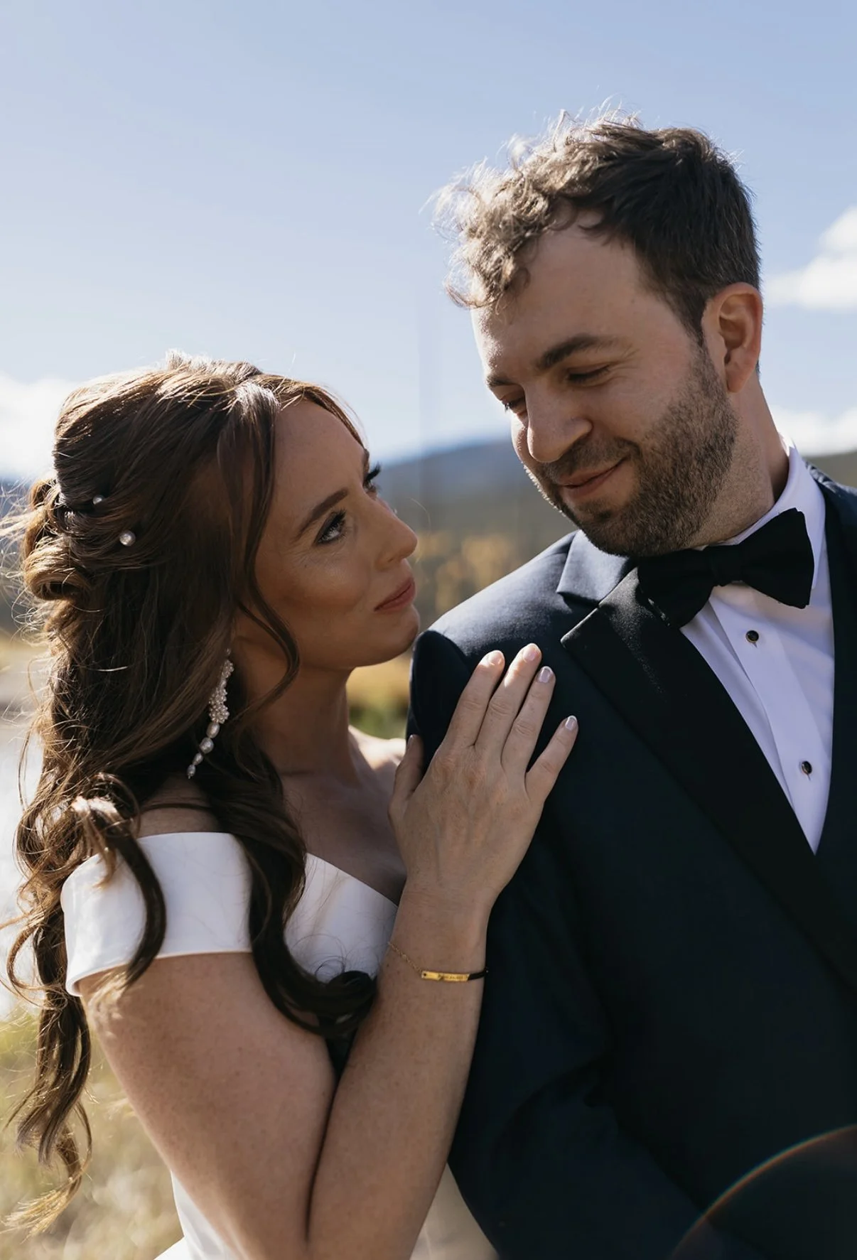 A woman with long brown curly hair and pearl earrings touching the shoulder of a man in a navy tuxedo, outdoors on a sunny day.