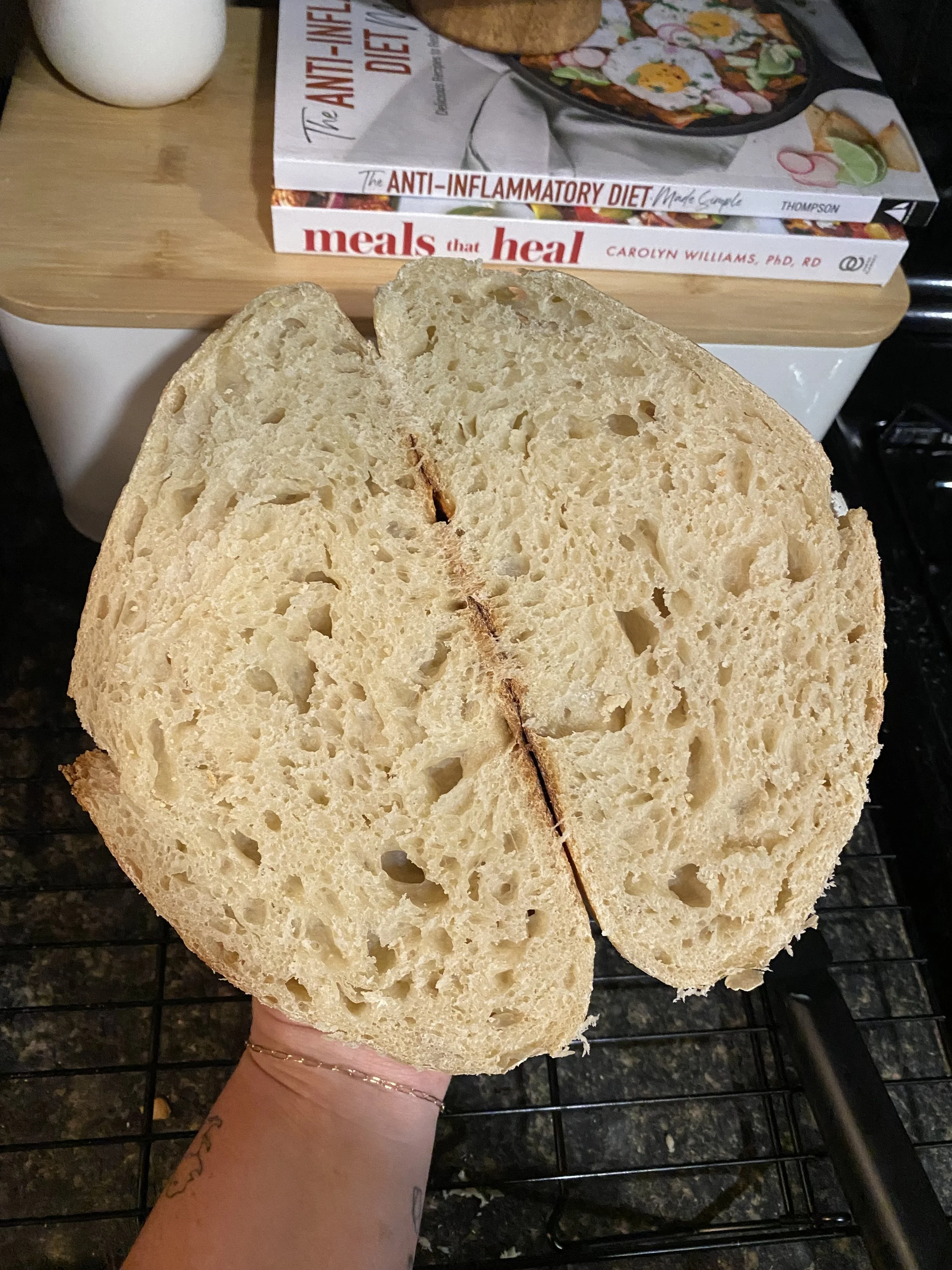 A person holding a large slice of bread with a hole in the middle. In the background, there are two books stacked on a wooden surface, with a white vase nearby. The books are titled 'The Anti-Inflammatory Diet Made Easy' and 'Meals That Heal'. The person’s wrist has a small tattoo and a gold bracelet.