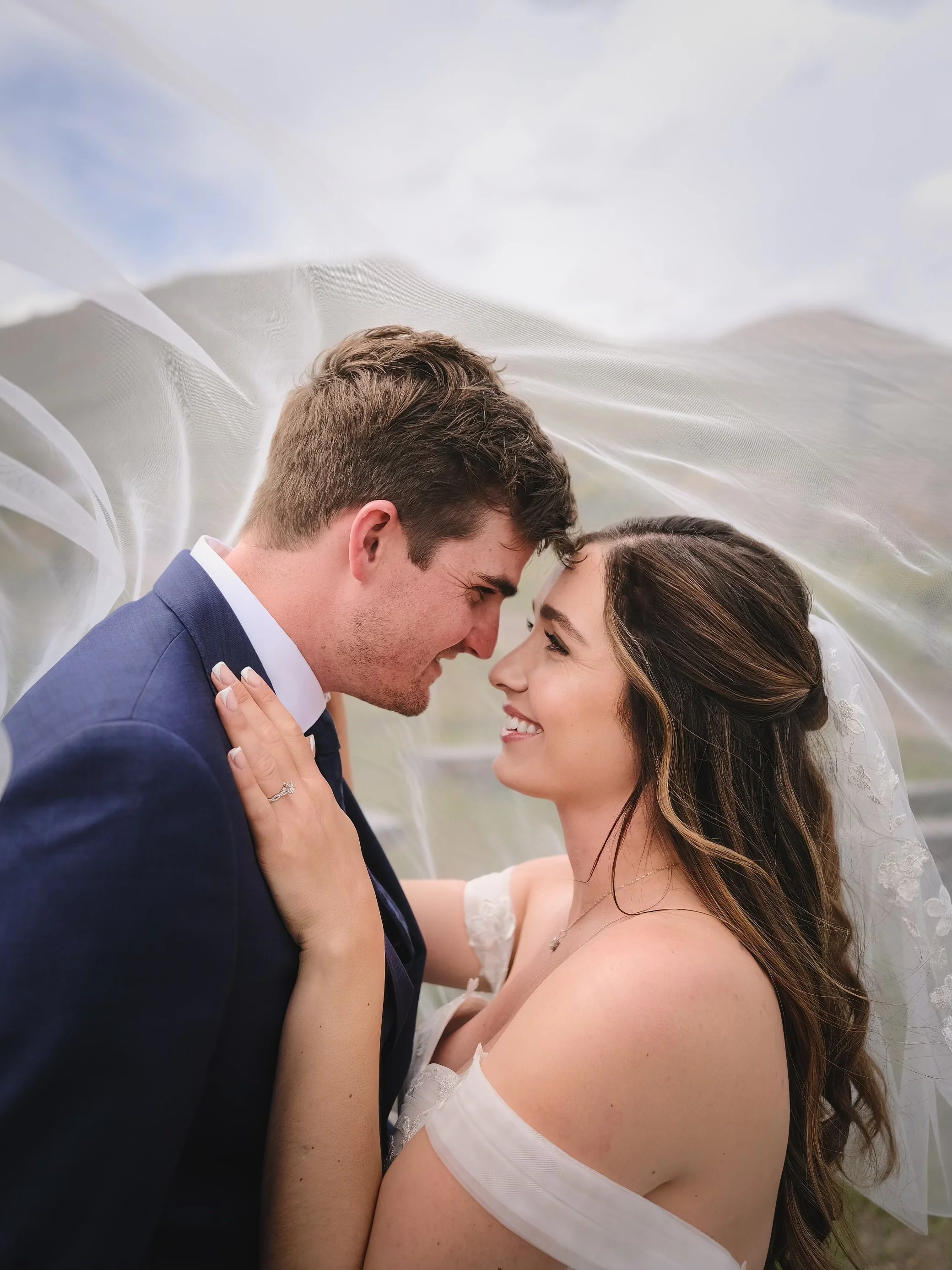 A bride and groom standing close together, touching foreheads and smiling, under a flowing white veil outdoors.