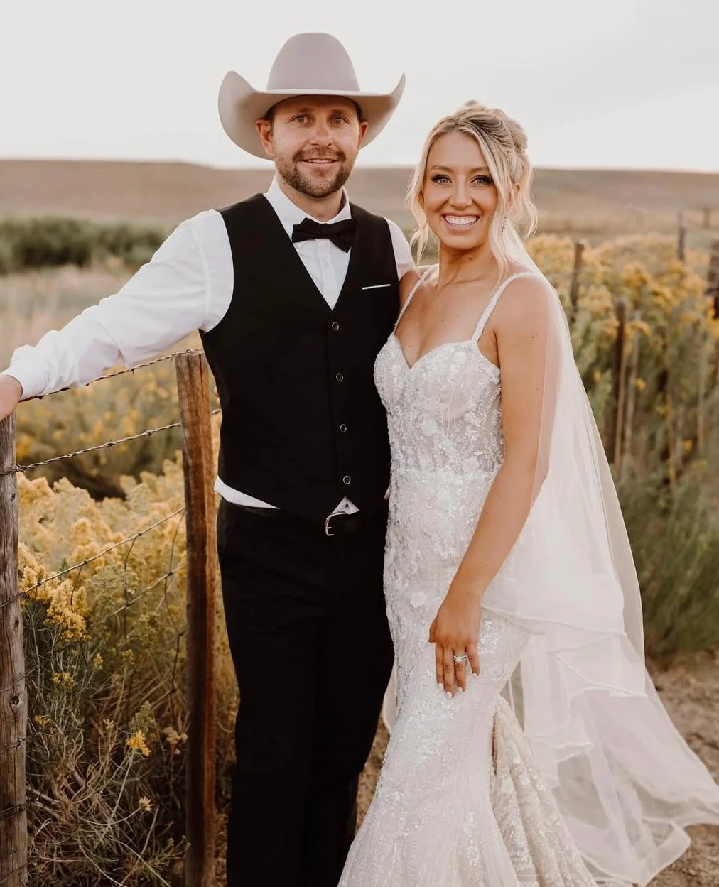 A newlywed couple smiling outdoors in a rural setting, with the groom wearing a cowboy hat, vest, and bow tie, and the bride in a lace wedding dress with a veil.