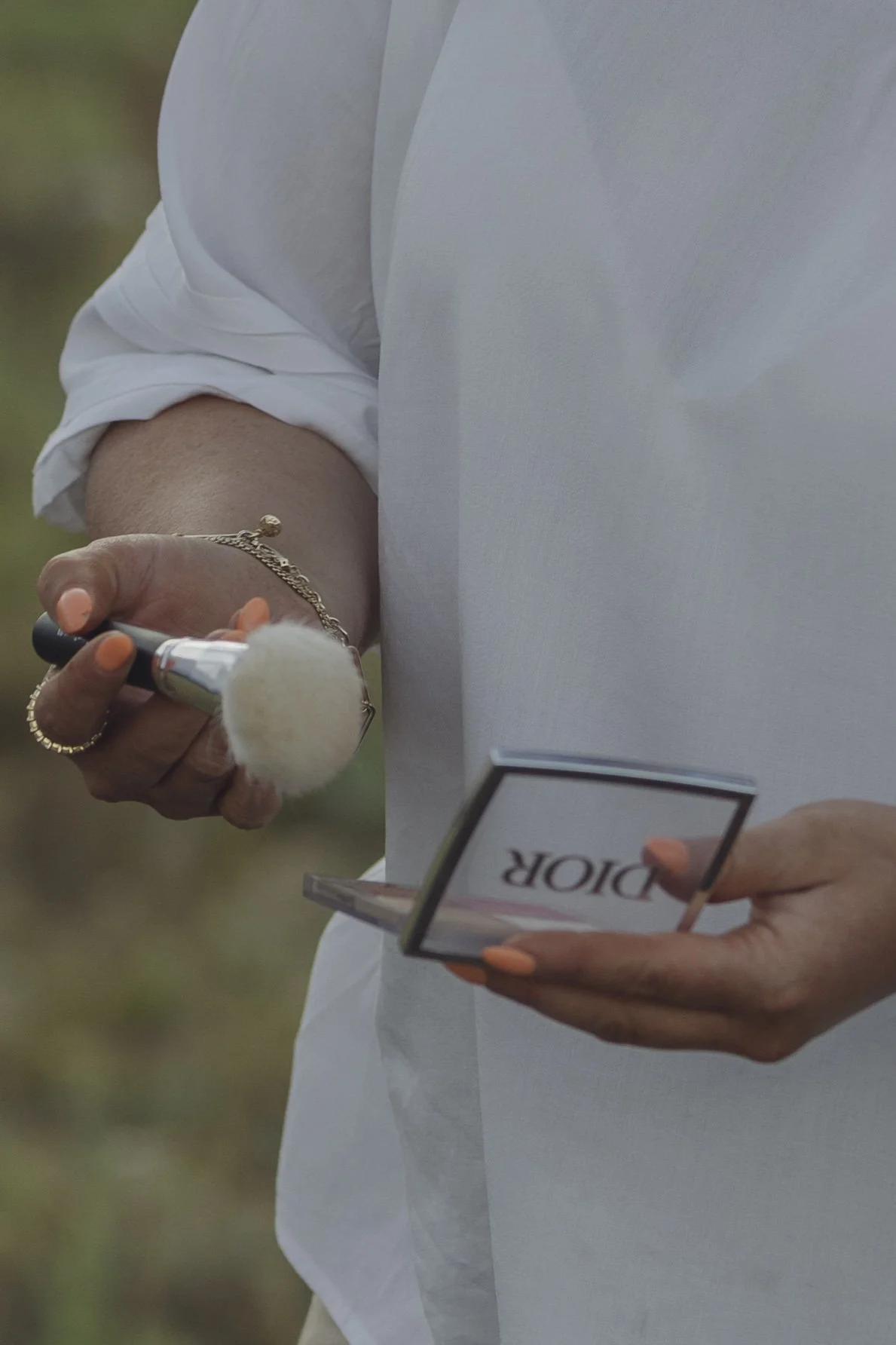Person holding a makeup brush in one hand and a Dior makeup palette in the other, wearing a white top, with a blurred green background.