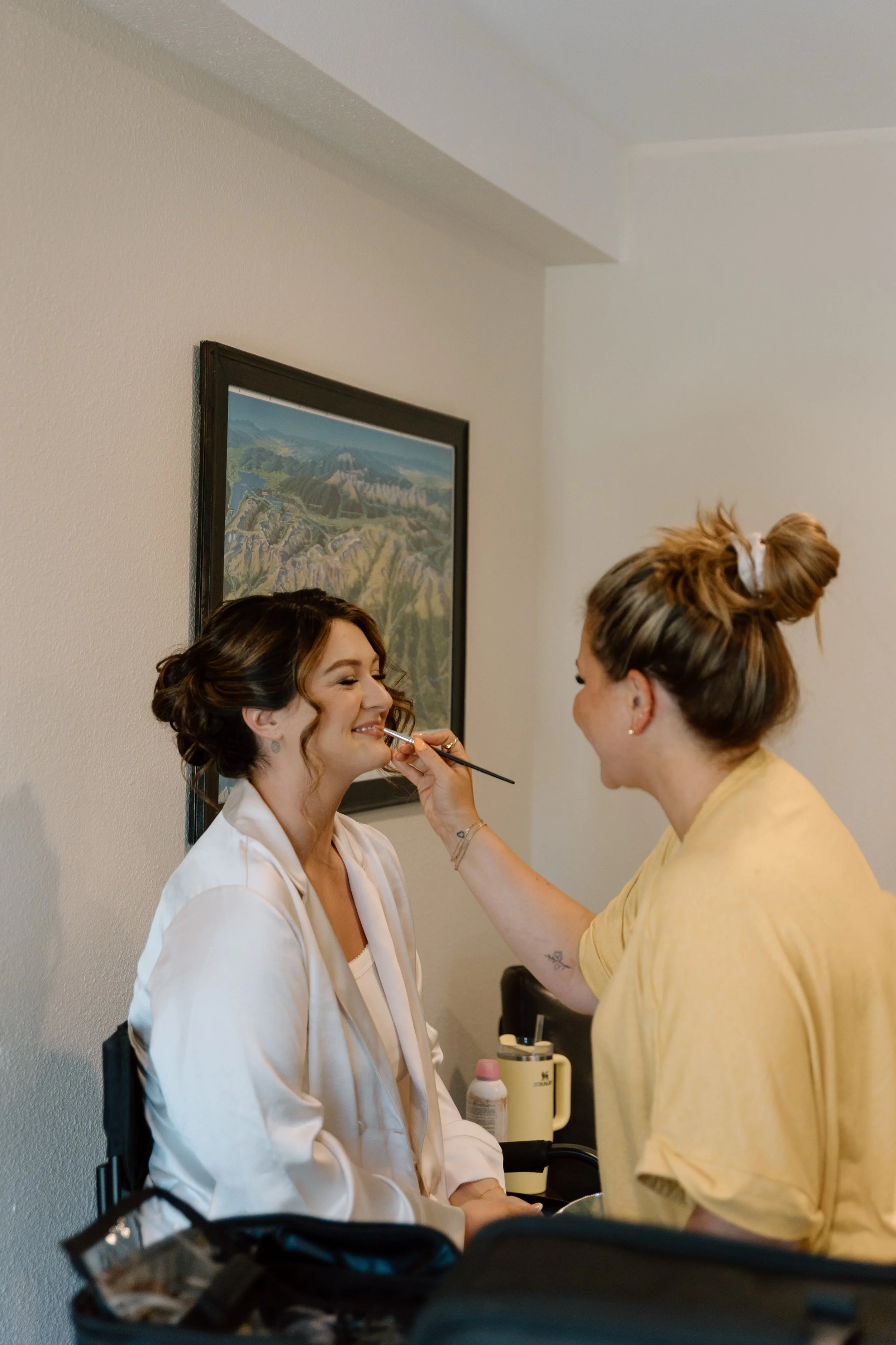 A makeup artist applies makeup to a woman with styled hair, who is seated and wearing a white robe. The setting appears to be a backstage or dressing room, with a framed landscape painting on the wall.
