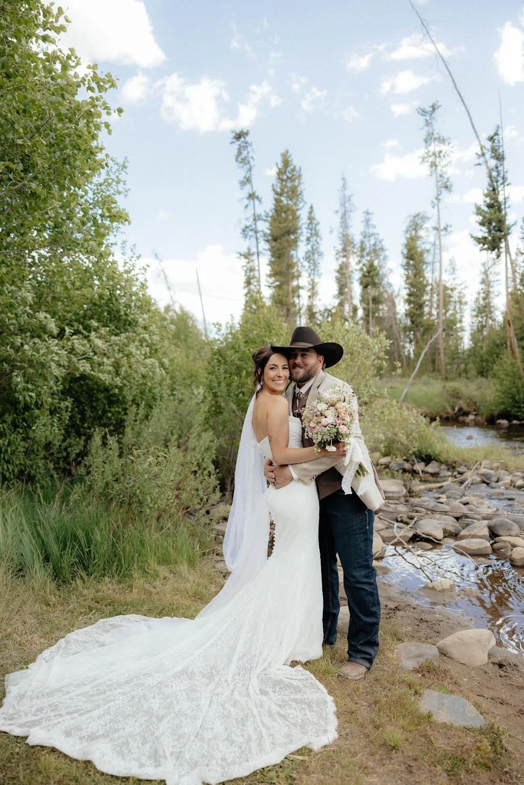 A newlywed couple stands outdoors near a river, smiling and embracing; the bride wears a white lace wedding gown with a long train, and the groom wears a cowboy hat, beige jacket, and dark jeans, holding a bouquet of flowers, with green trees and a blue sky in the background.