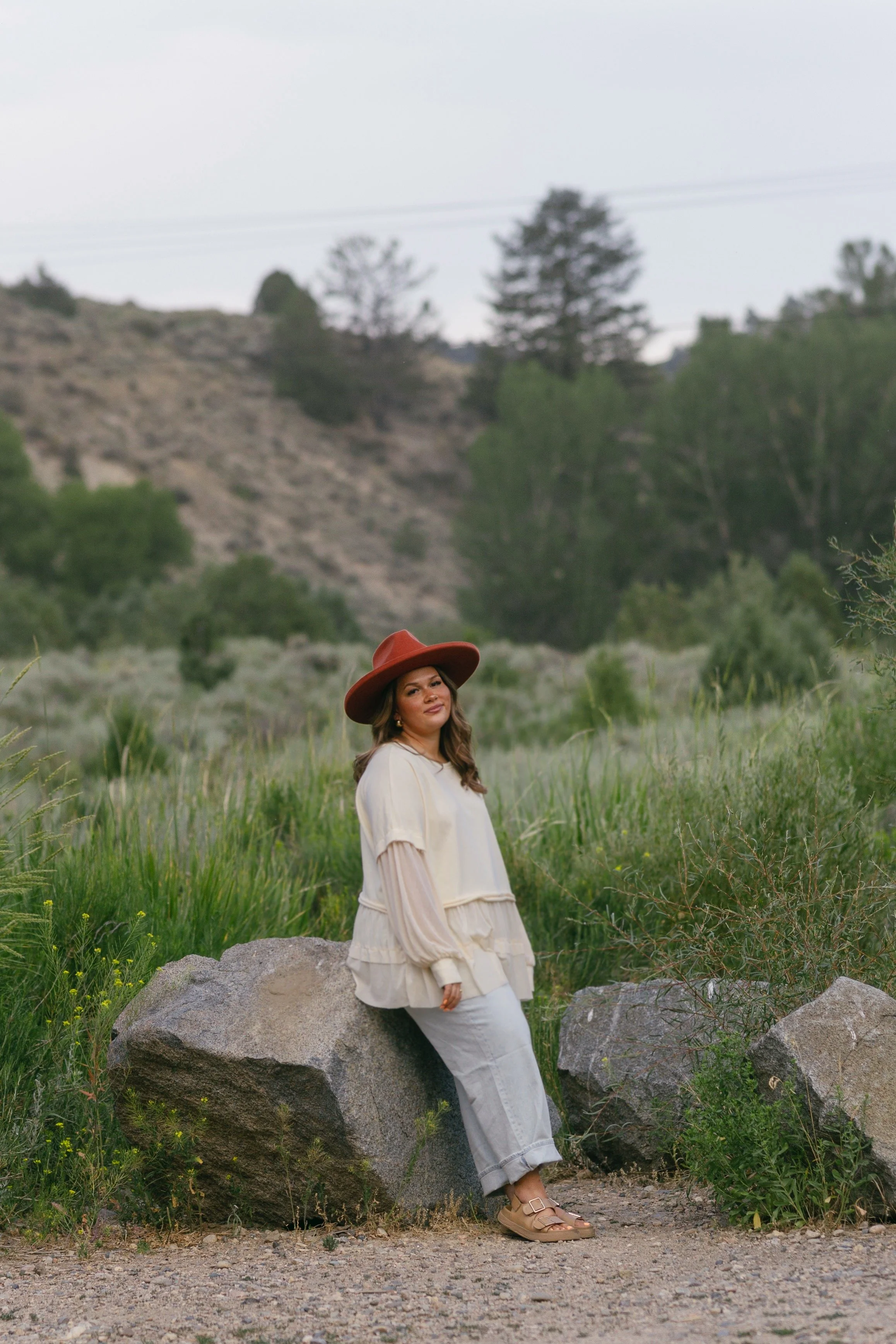 A woman wearing a beige dress and a large brown hat standing next to a large rock in a green, hilly outdoor landscape with trees and mountains in the background.