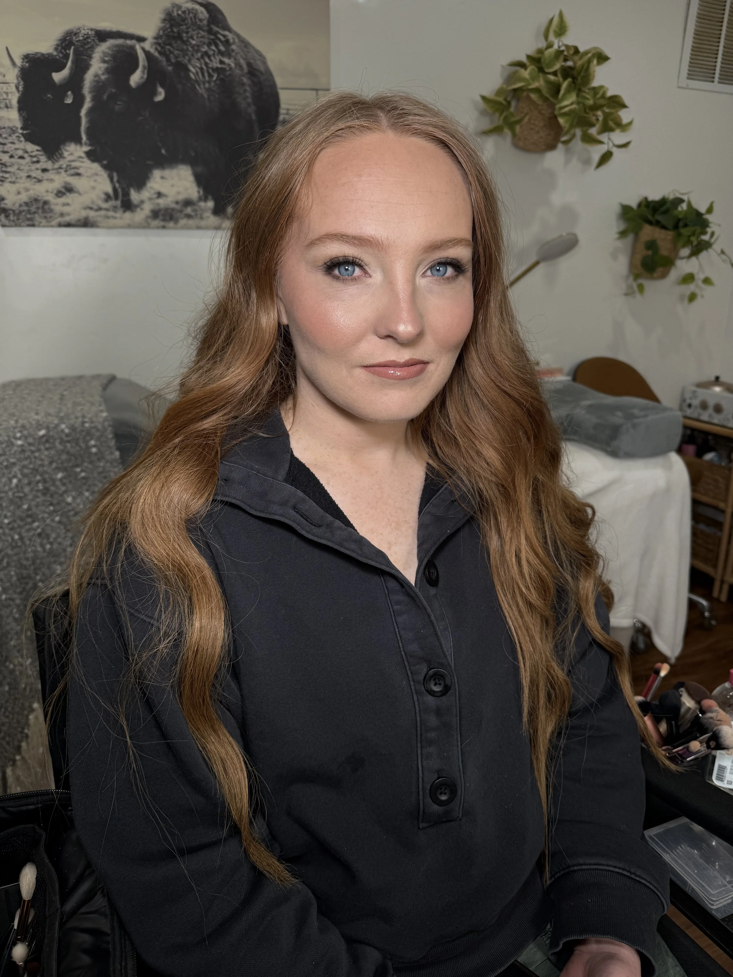 A woman with long red hair and blue eyes sitting in front of a wall with a black and white buffalo poster, plants, and makeup tools in the background.