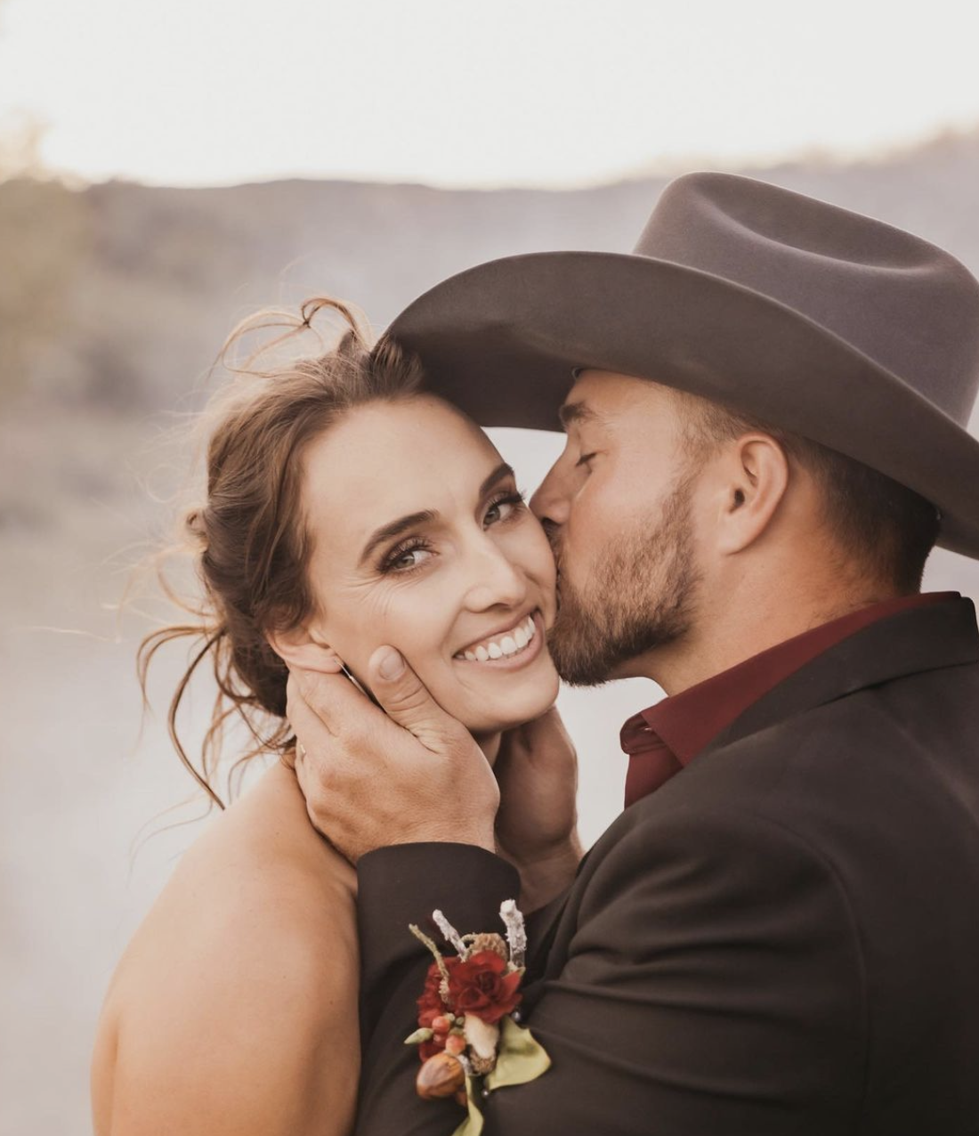 A couple sharing a kiss outdoors; the woman is smiling at the camera while embracing the man, who is kissing her cheek. The man is wearing a large cowboy hat and a dark suit with a red shirt, and the woman has her hair in an updo, with a floral corsage on her wrist.