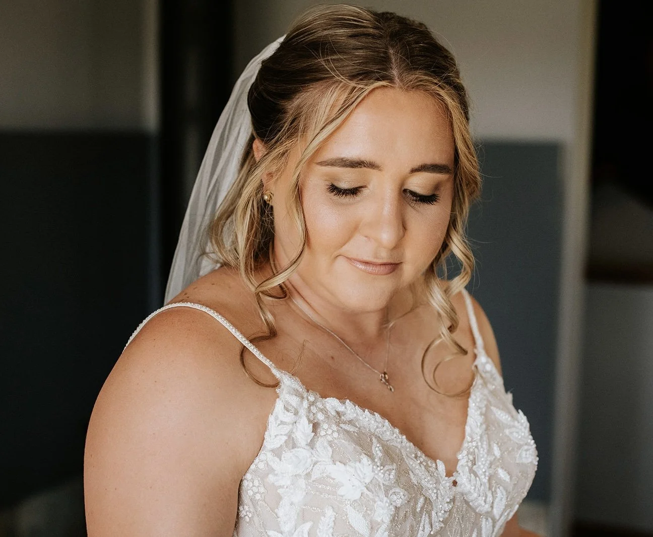 A bride with blonde curls and a veil, wearing a white lace wedding dress, with her eyes closed and a peaceful expression.