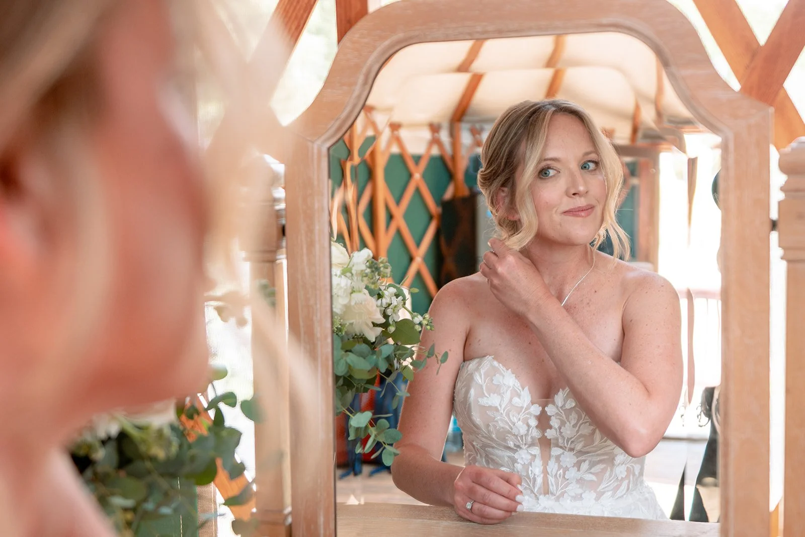 A woman dressed in a wedding gown looking at herself in a mirror as she adjusts her hair.