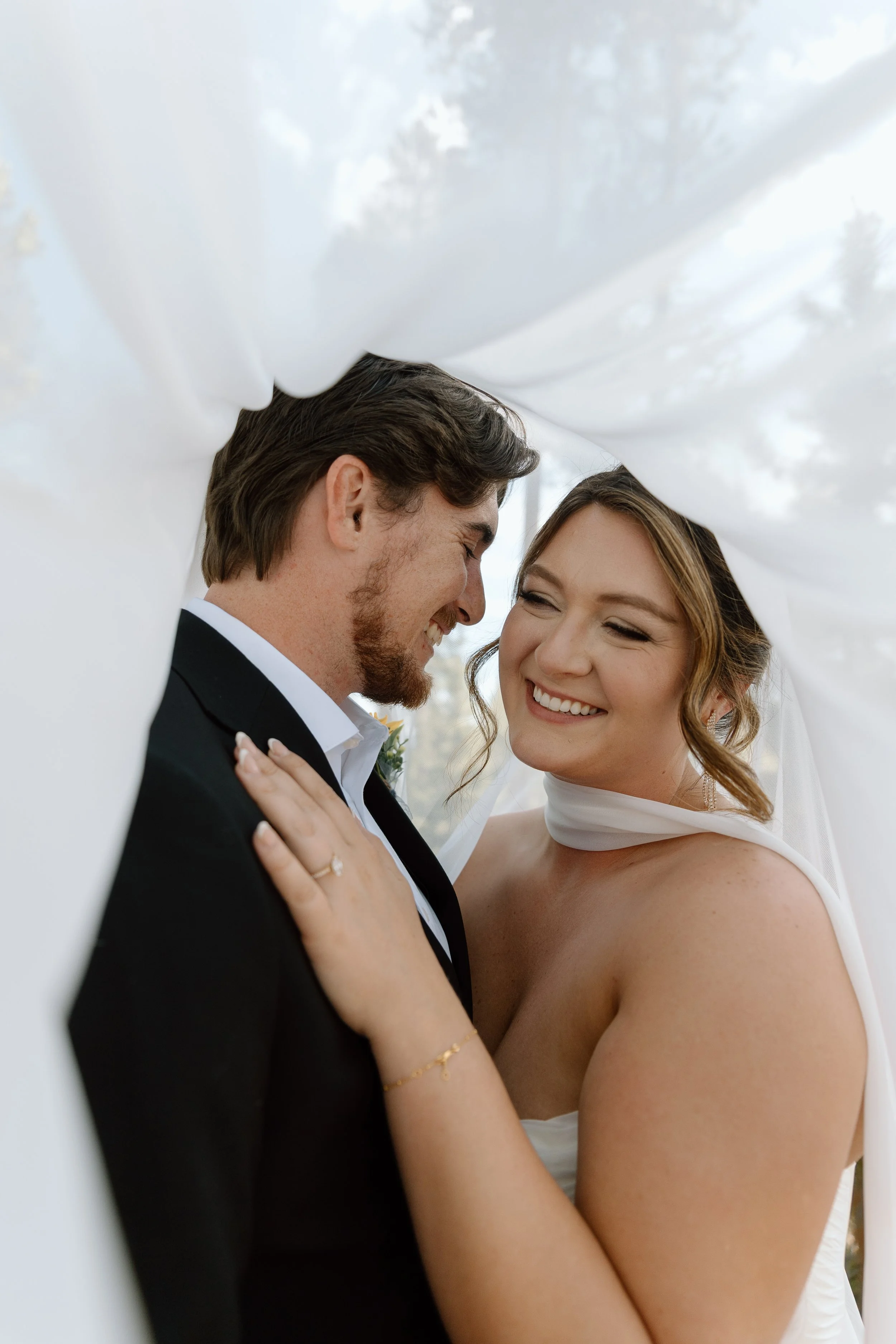 A newlywed couple smiling and sharing an intimate moment outdoors, with white fabric and cloudy sky in the background.
