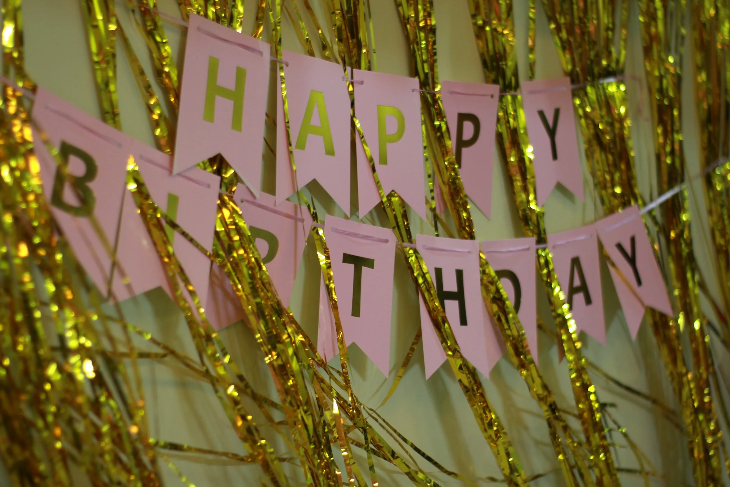 Pink and gold 'Happy Birthday' banner hanging against a shiny gold streamers background.