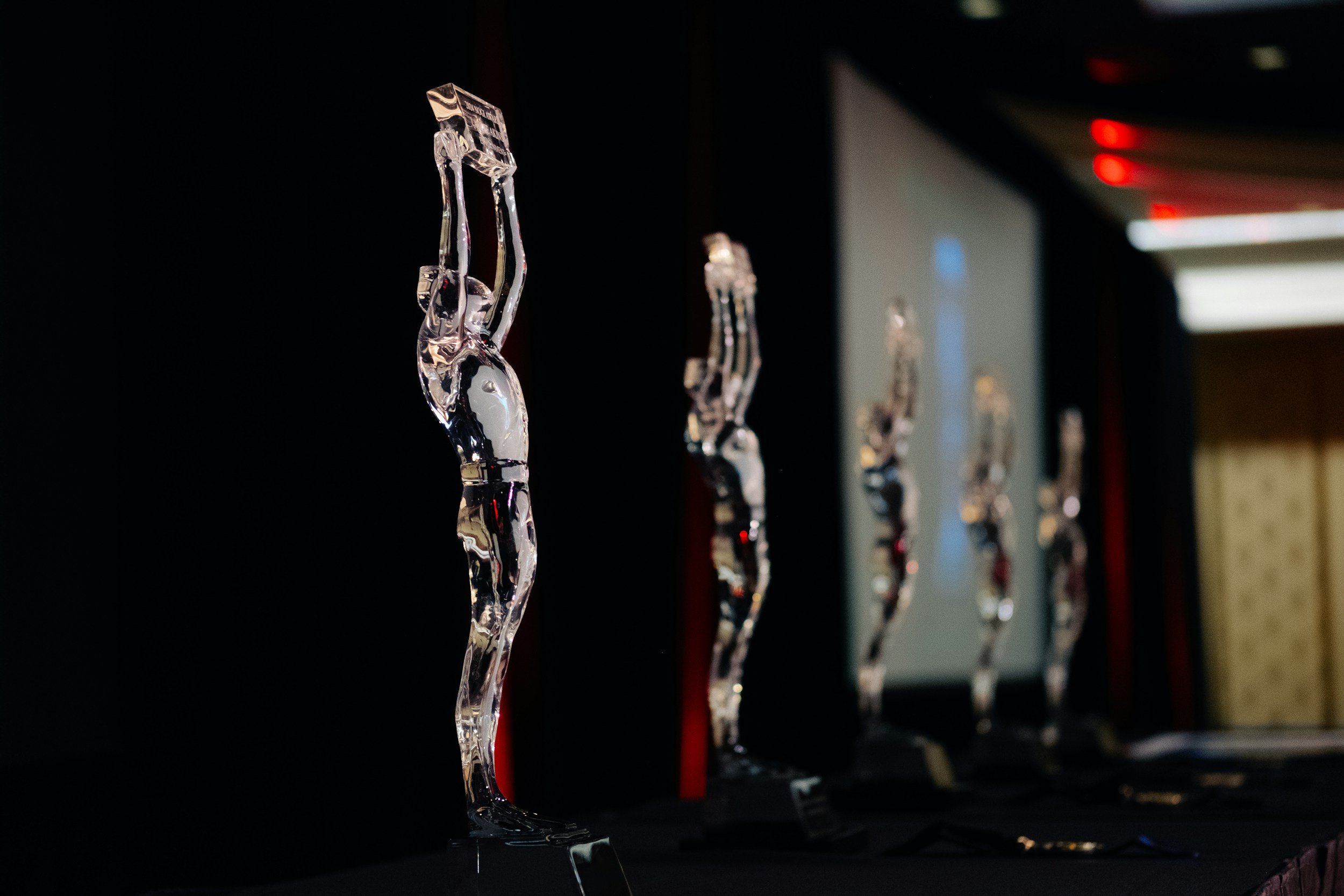 Close-up of a clear glass award or trophy shaped like a figure with an arm raised, displayed on a black surface against a dark background, with other similar awards blurred in the background.