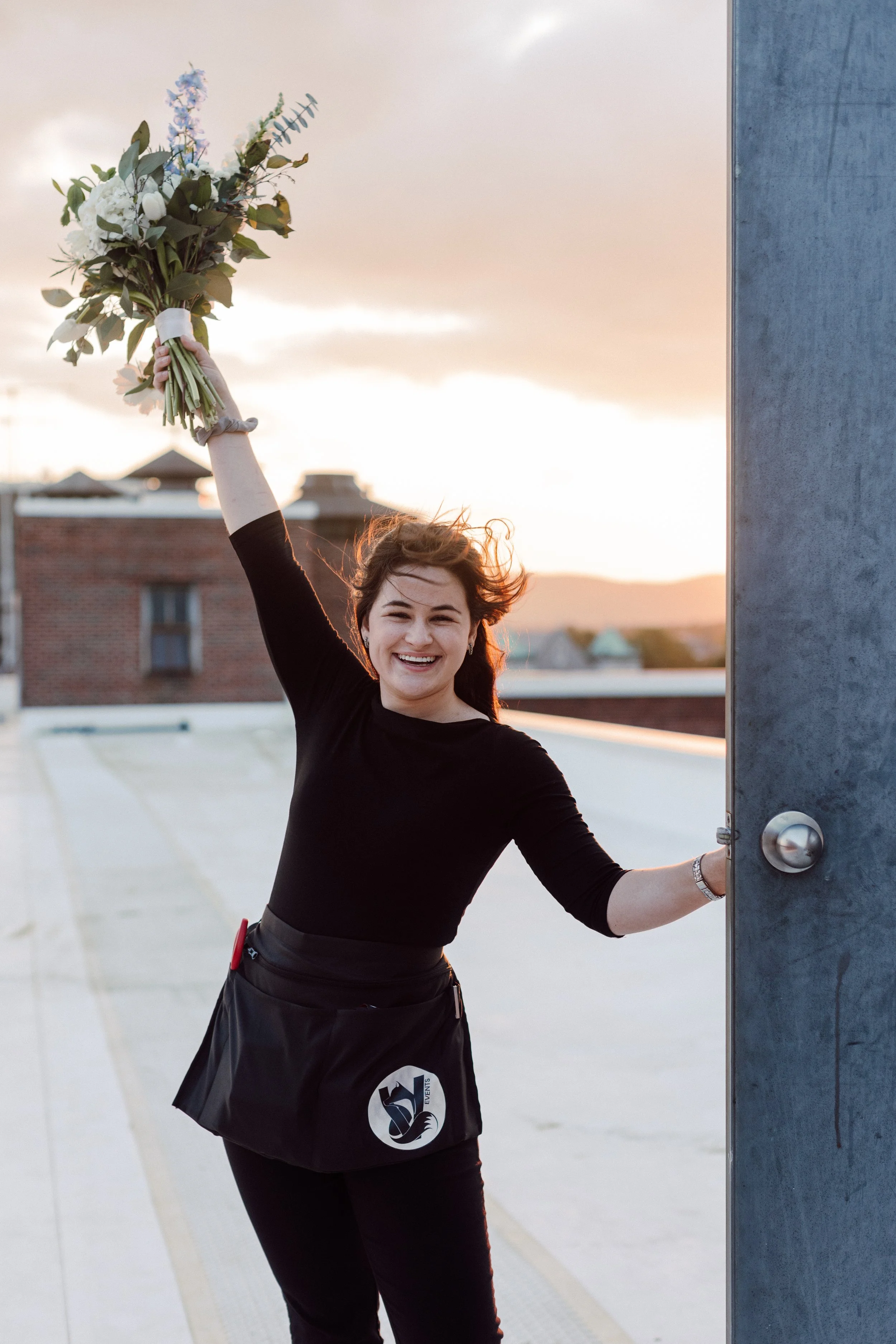 A smiling woman with brown hair in a black long-sleeve shirt and black skirt stands on a rooftop at sunset, holding a large bouquet of flowers high in her right hand and the rooftop door with her left hand.