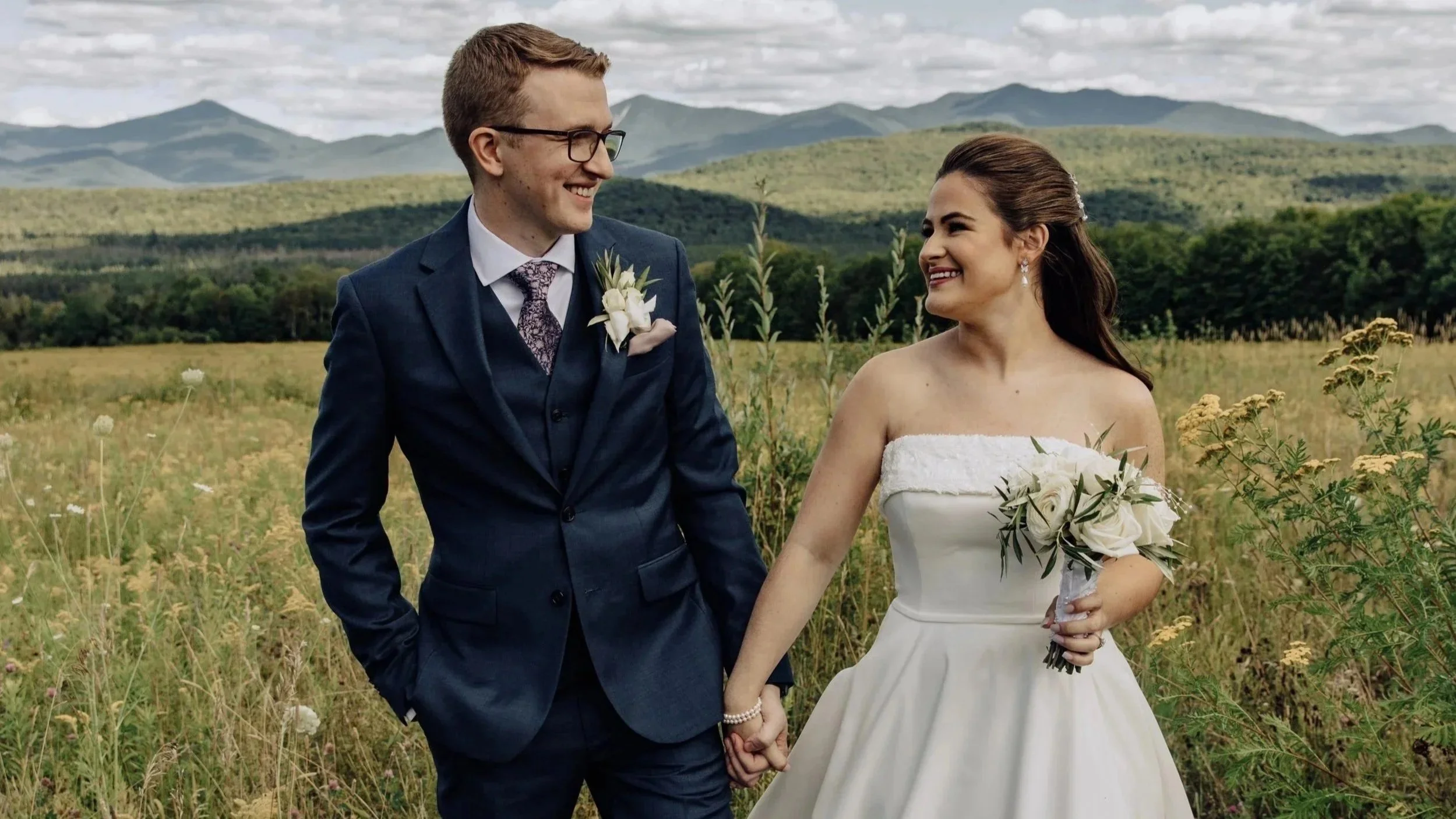 A newlywed couple holding hands and smiling in a field of wildflowers with adirondack mountains in the background.