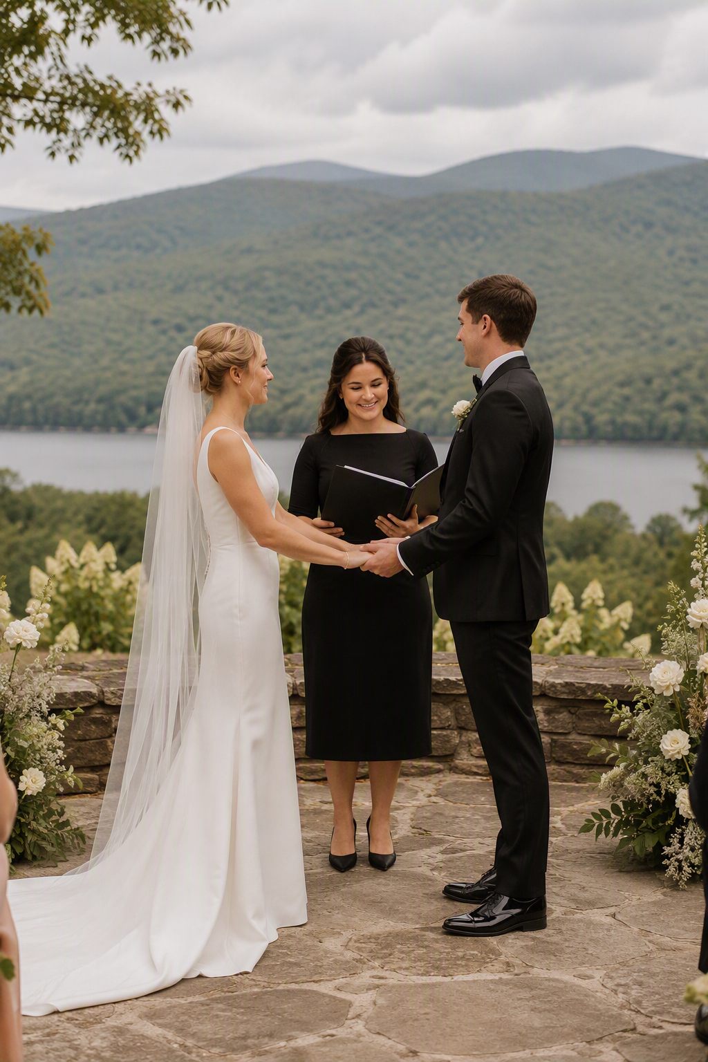 A bride and groom holding hands during their wedding ceremony outdoors, with a lake and mountain landscape in the background, officiant standing between them, flowers decorating the ceremony site.