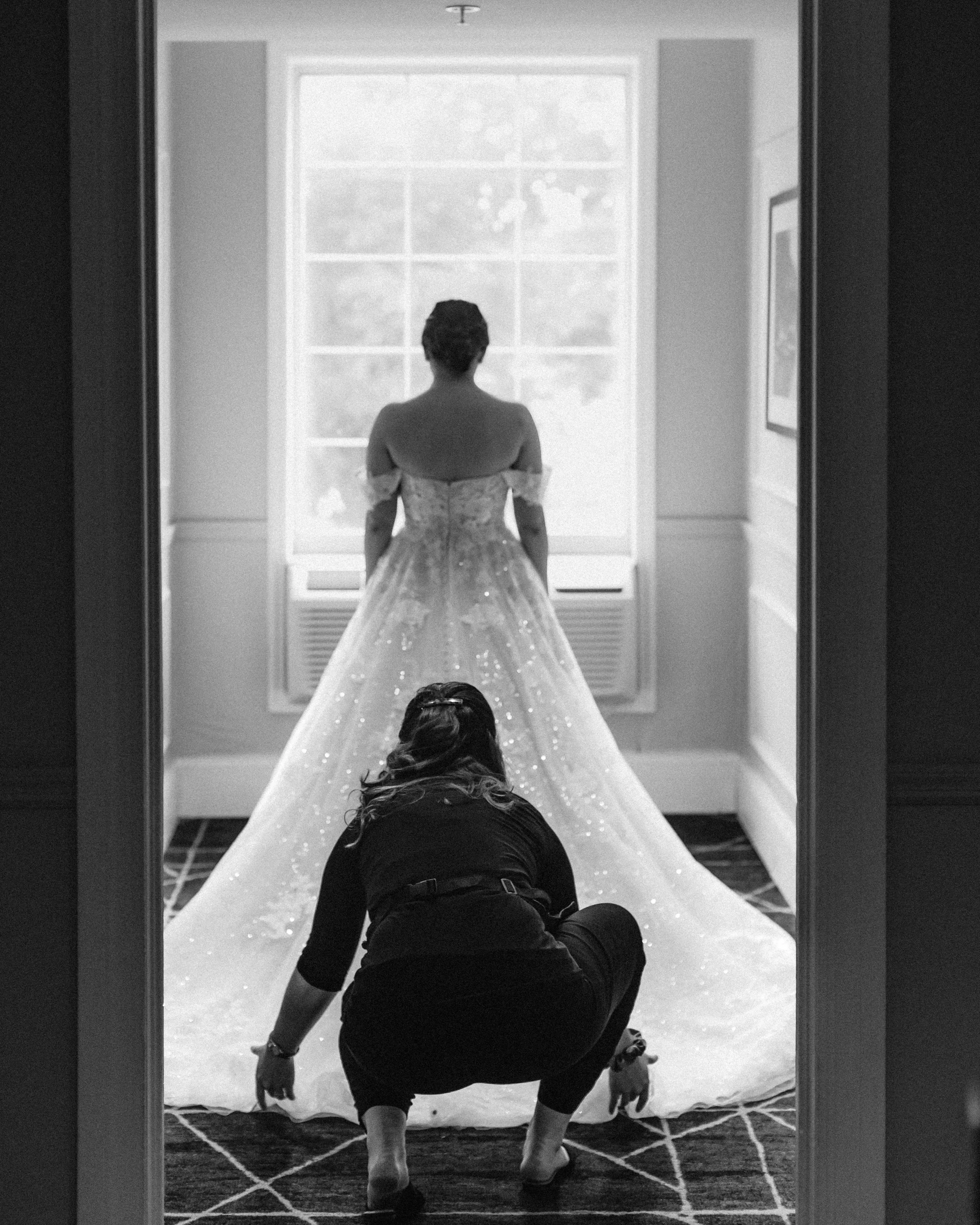 A person kneeling on the floor in front of a large mirror, adjusting a wedding dress on someone sitting in front of the mirror. The person in the dress has their back to the camera, and the scene is lit with natural light coming through a window in the background.