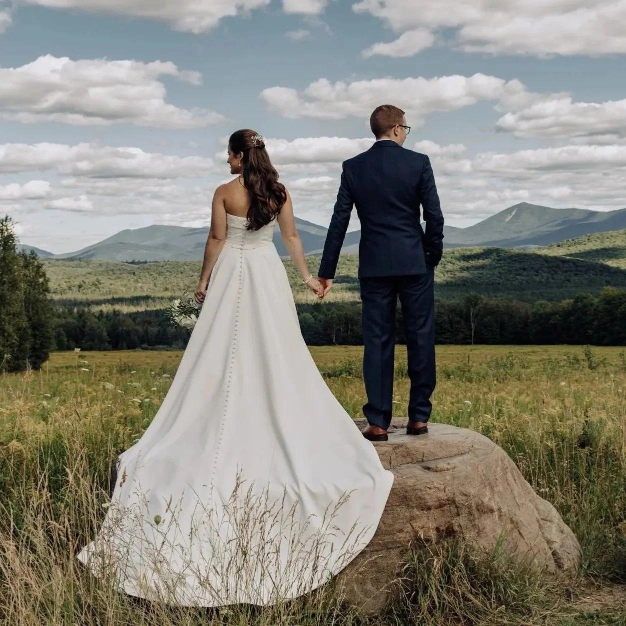 A bride and groom holding hands on a large rock in a grassy field with mountains and a cloudy sky in the background.