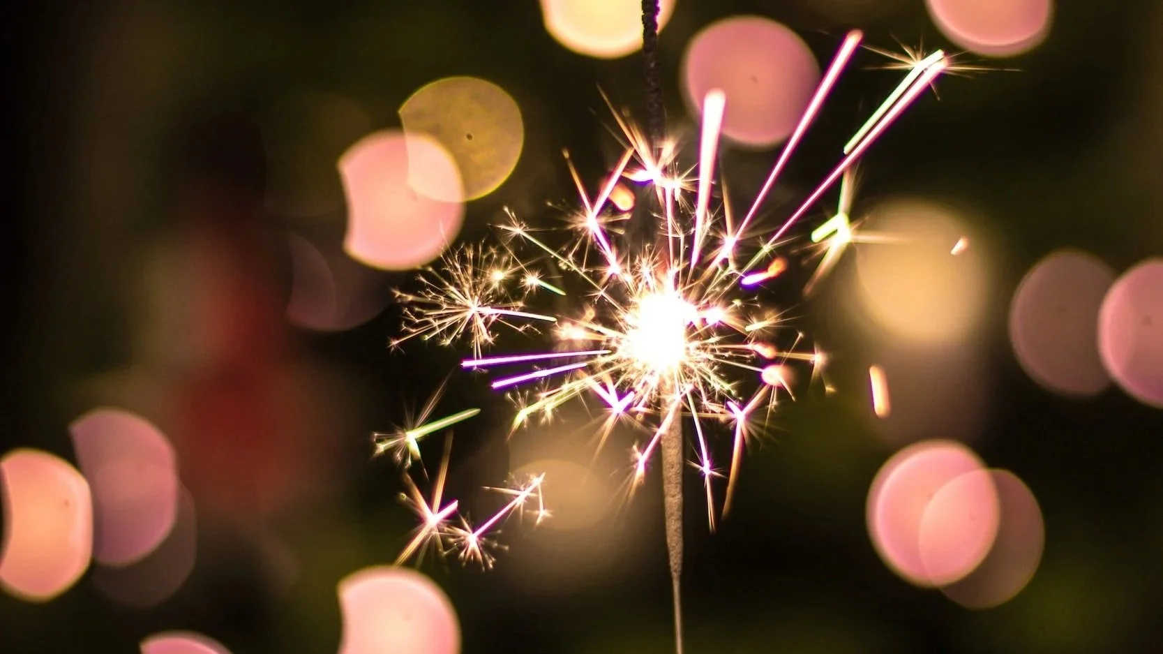 A close-up of a lit sparkler emitting bright sparks against a dark background with pink and gold  lights.