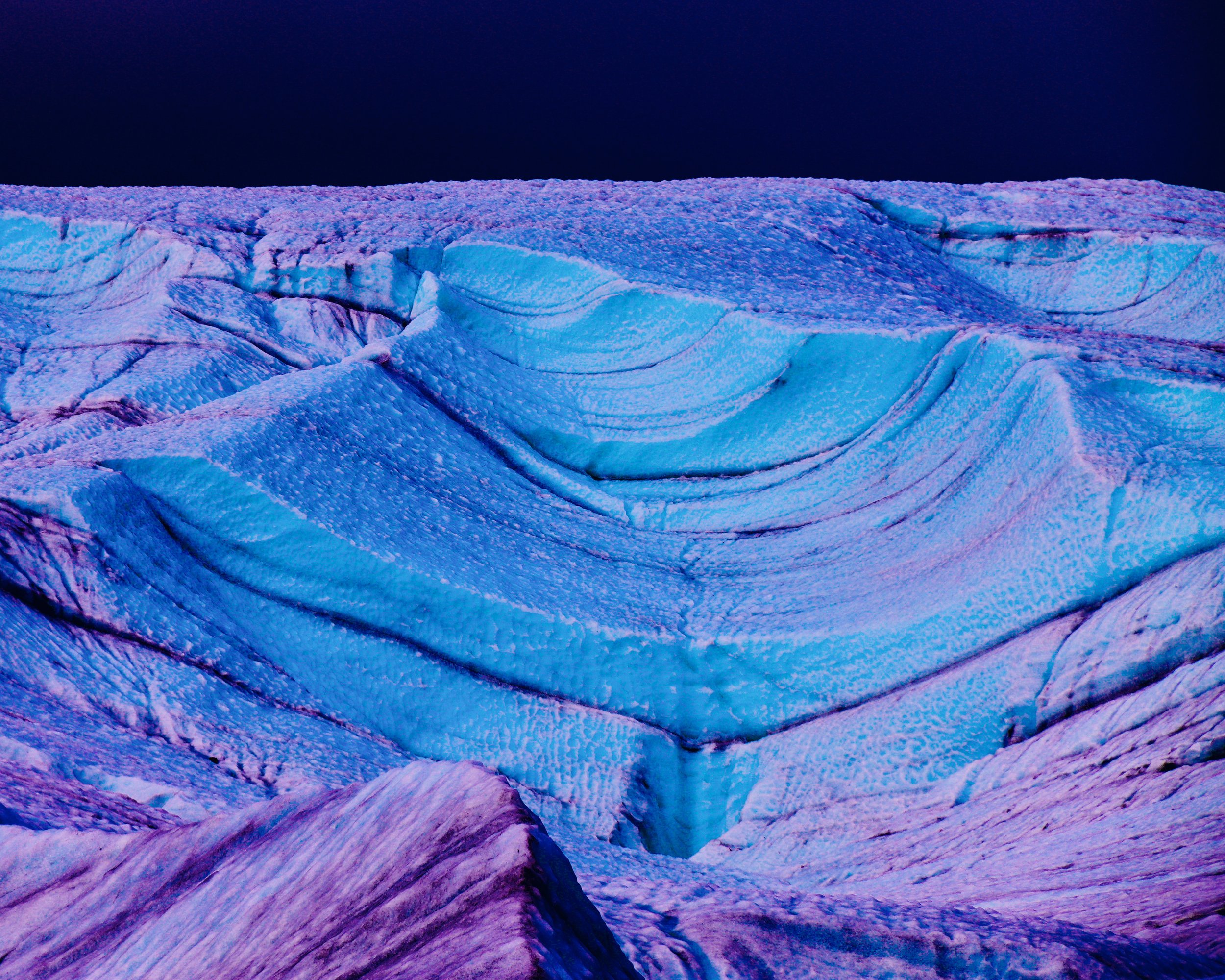 Nighttime view of a glacier with colorful lighting highlighting the ice's textures and crevasses.