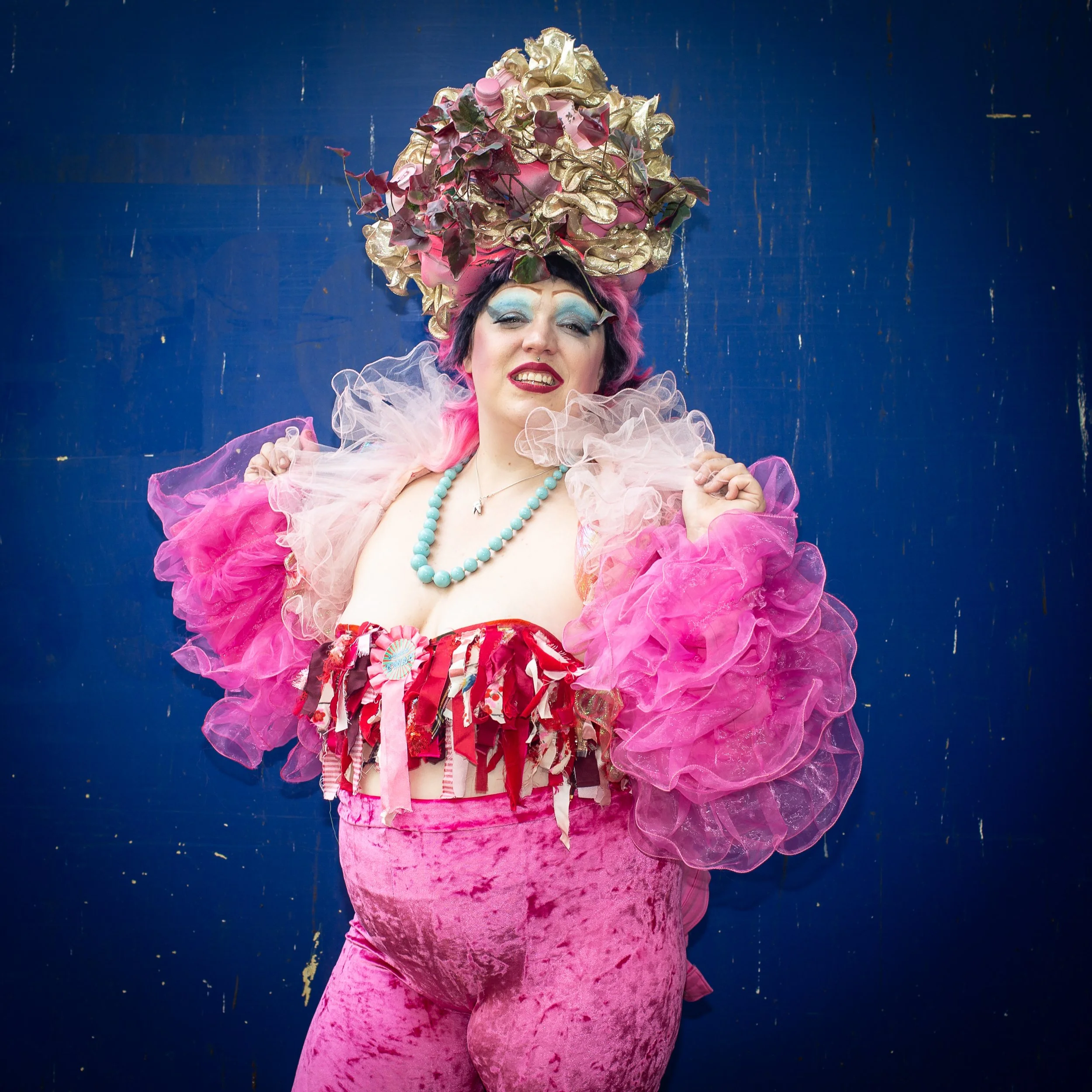 Clunge mother is standing in front of a blue background wearing pink and gold headpiece made out of plastic bottles. pink frilled jacket, pink velour leggings and bodice made of pink and red rags