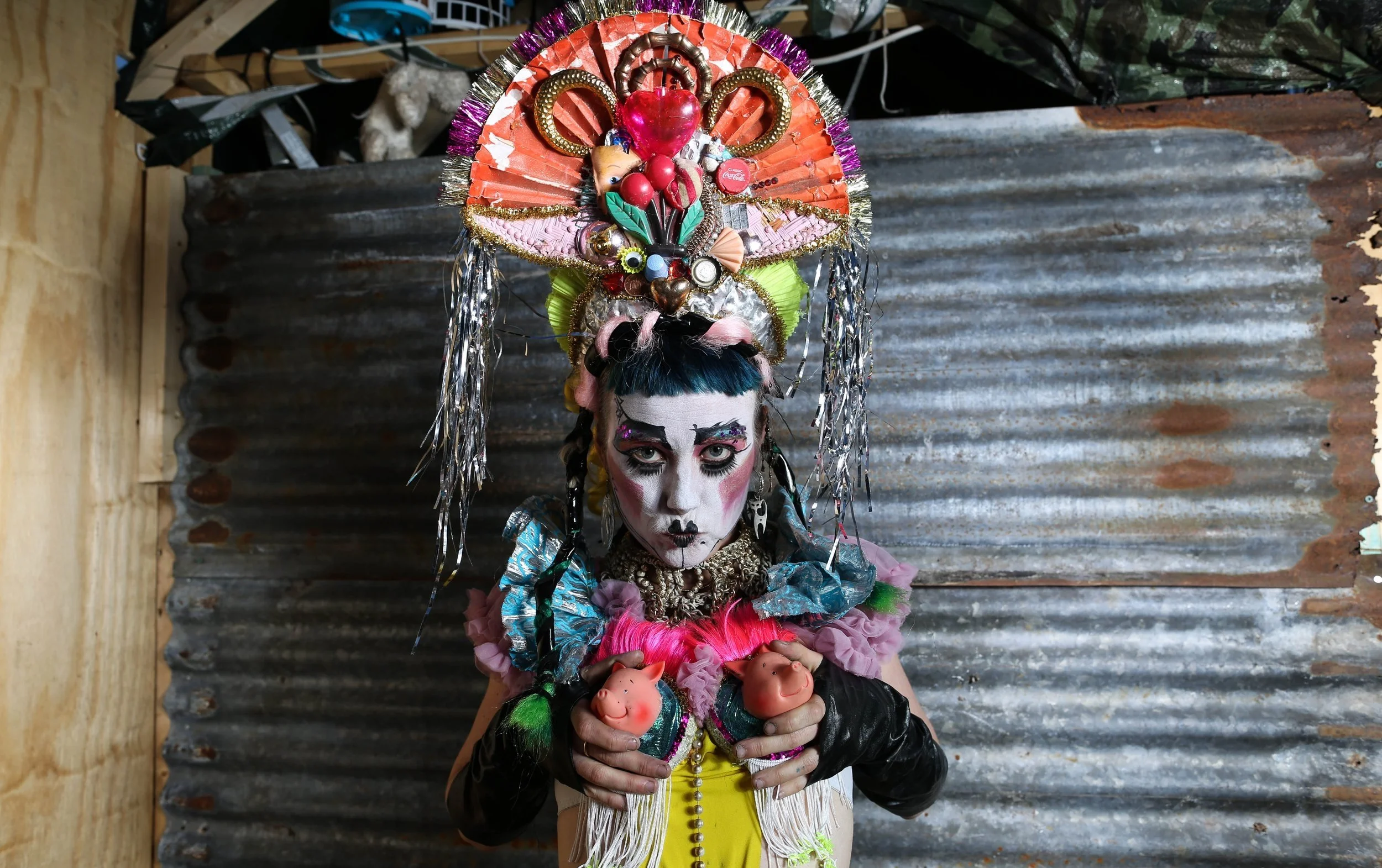 female performer in front of corrugated iron back drop. Wearing headpiece made from  an old orange fan, found objects, rubbish and tinsel, she has pig tails and a short blue fringe, make up is a white face and black cupid lips.