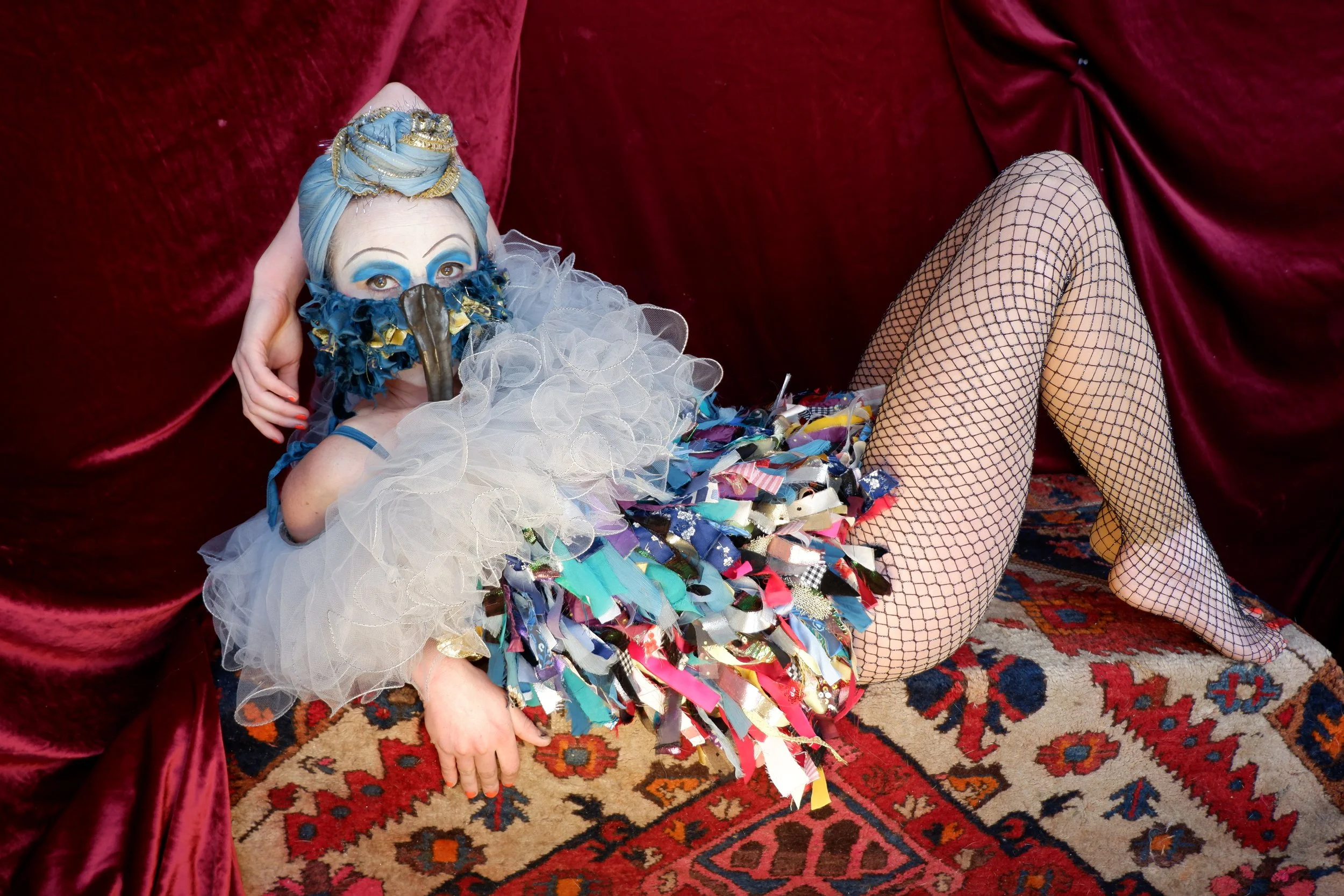Performer lounging on kilim carpet in front of red velvet curtain wearing bird circus costume, frills, bird mask, blue eyeshadow and blue and gold turban
