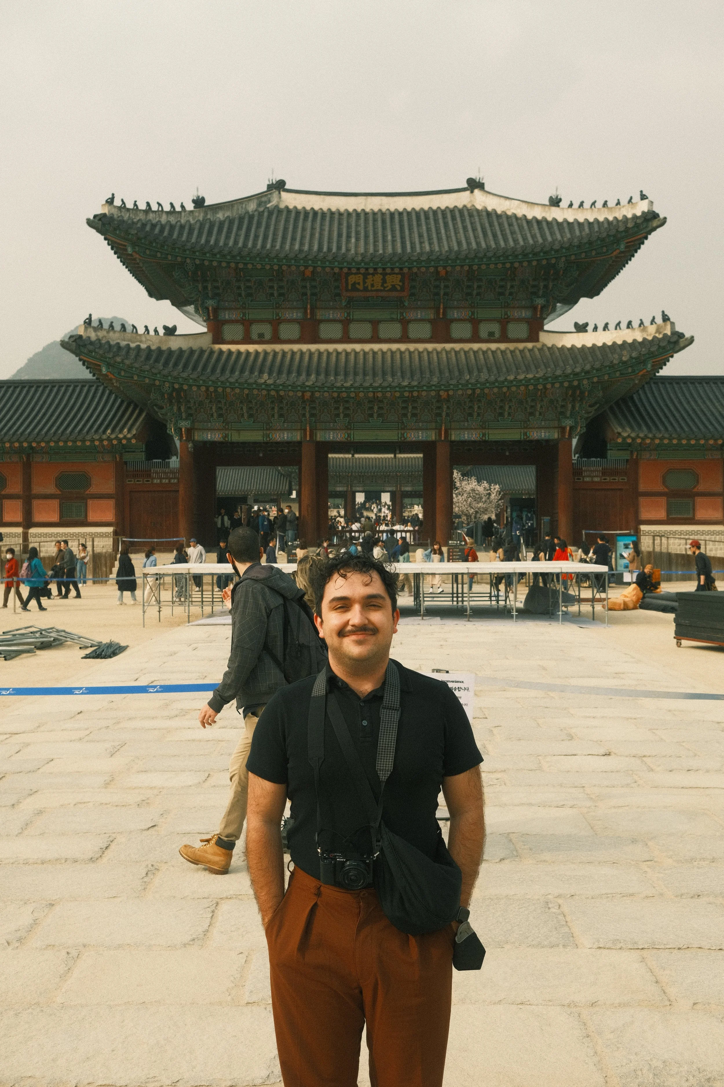 A man with a camera around his neck smiling in front of a traditional Asian temple with multiple tiered roofs and a large gate. There are people walking and happening in the background.