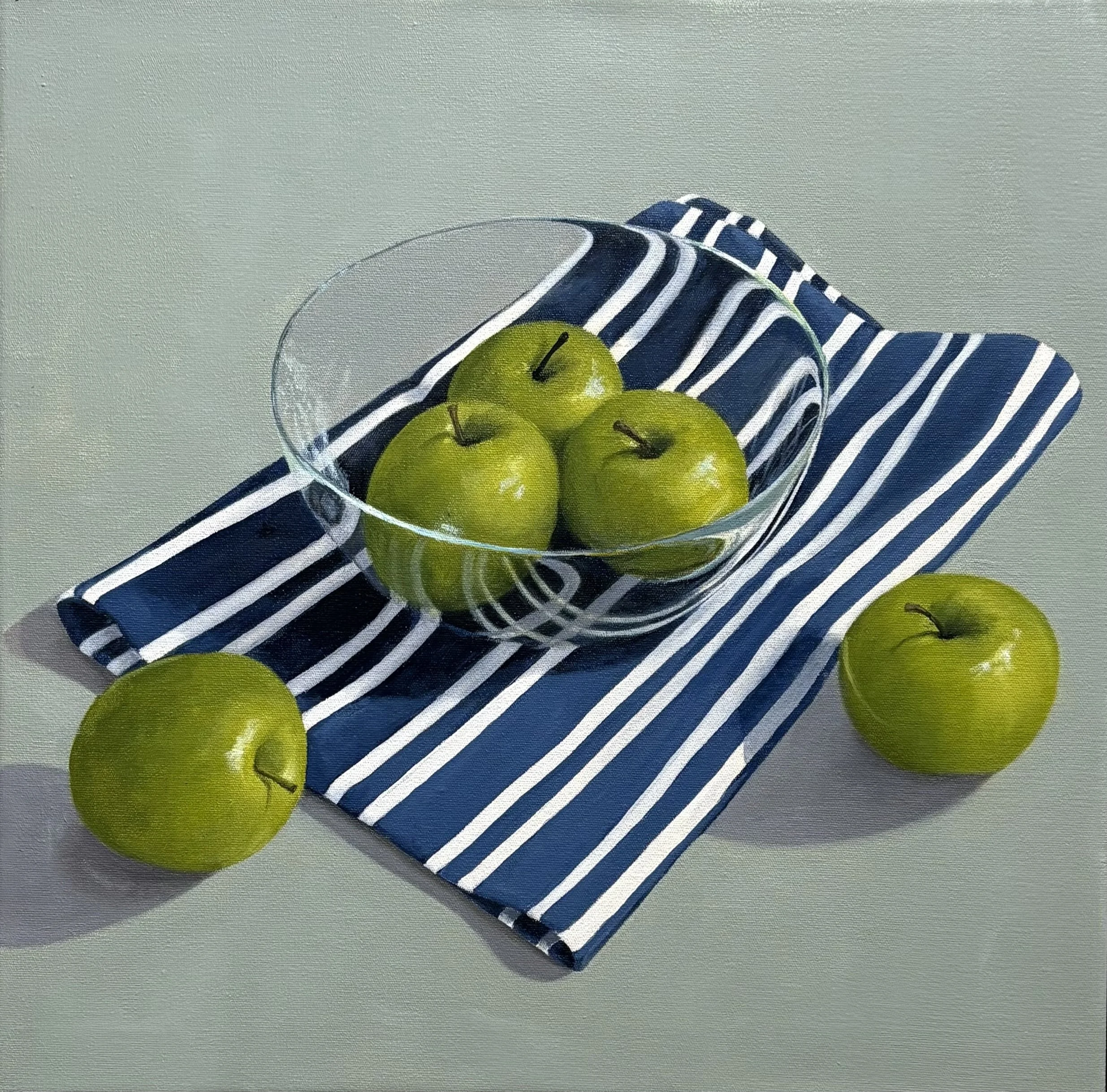 Green apples in a glass bowl on a blue and white striped cloth, with two additional apples outside the bowl on a light-colored surface.