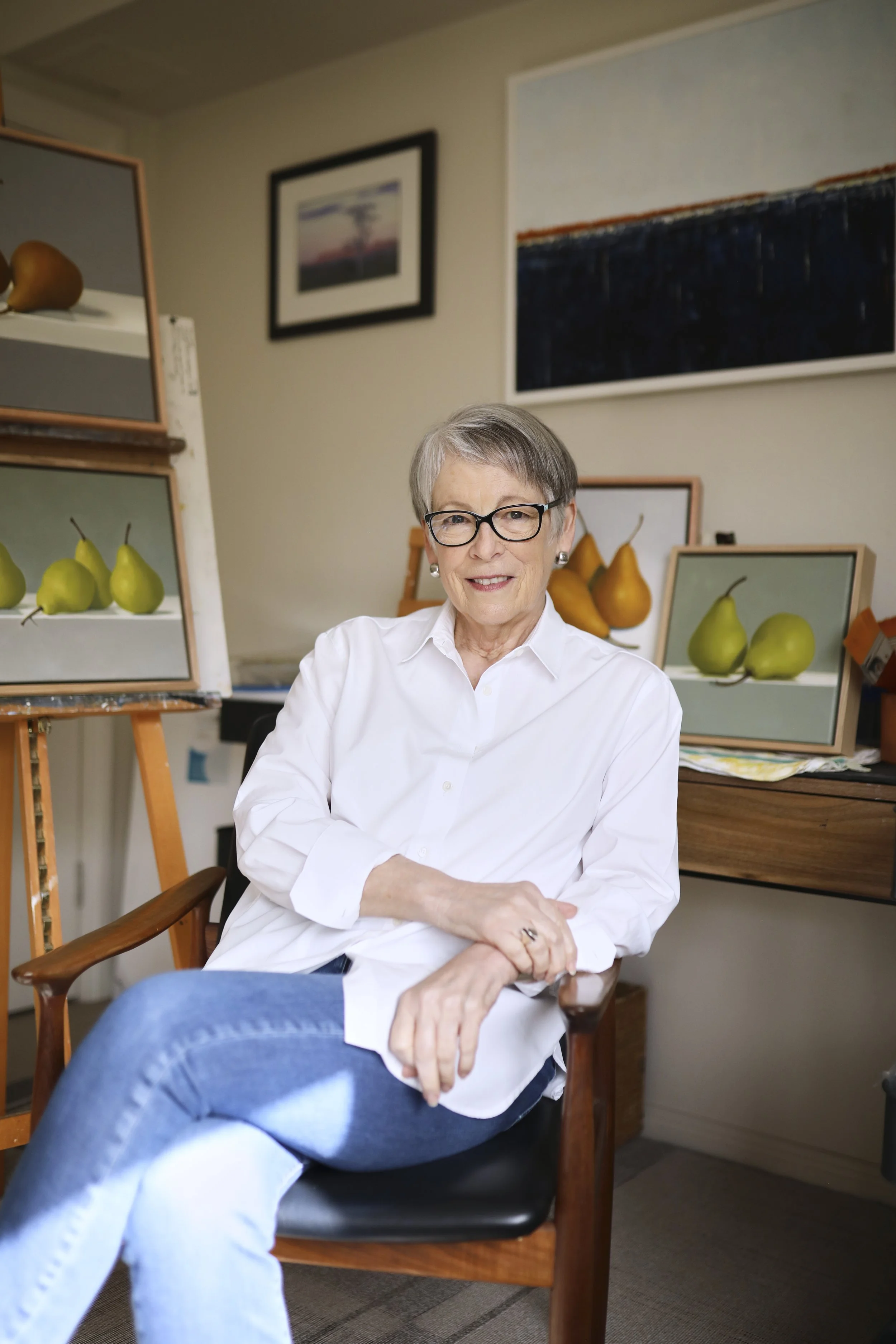 An artist, wearing a white shirt and jeans, sitting in a black and wooden chair in an art studio. She is surrounded by paintings, with her hands resting on her lap and smiling at the camera.