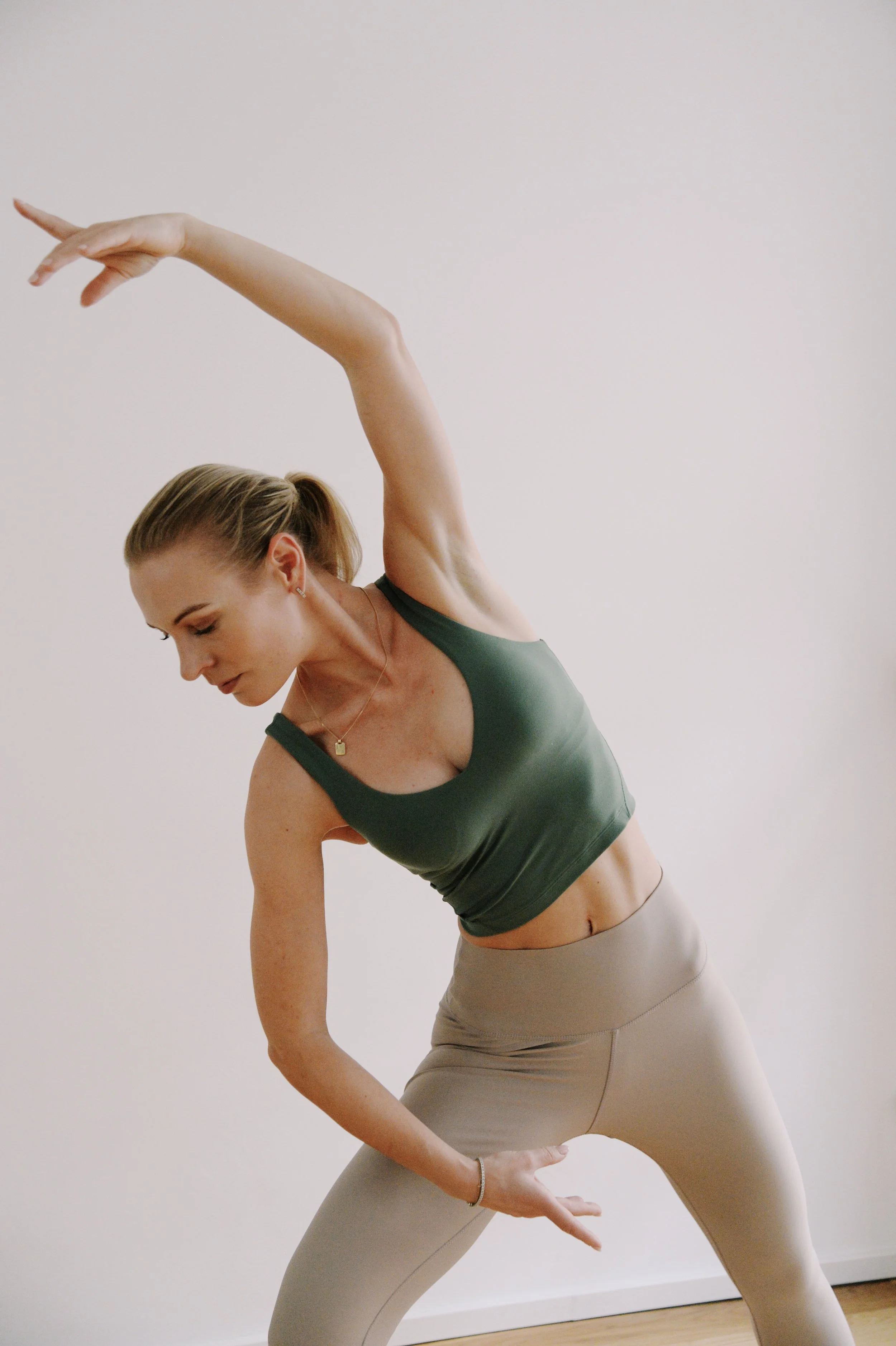 Eine Frau macht Yoga vor einer weißen Wand, trägt ein grünes Top und beige Leggings, die in einer Yoga-Pose stehen.