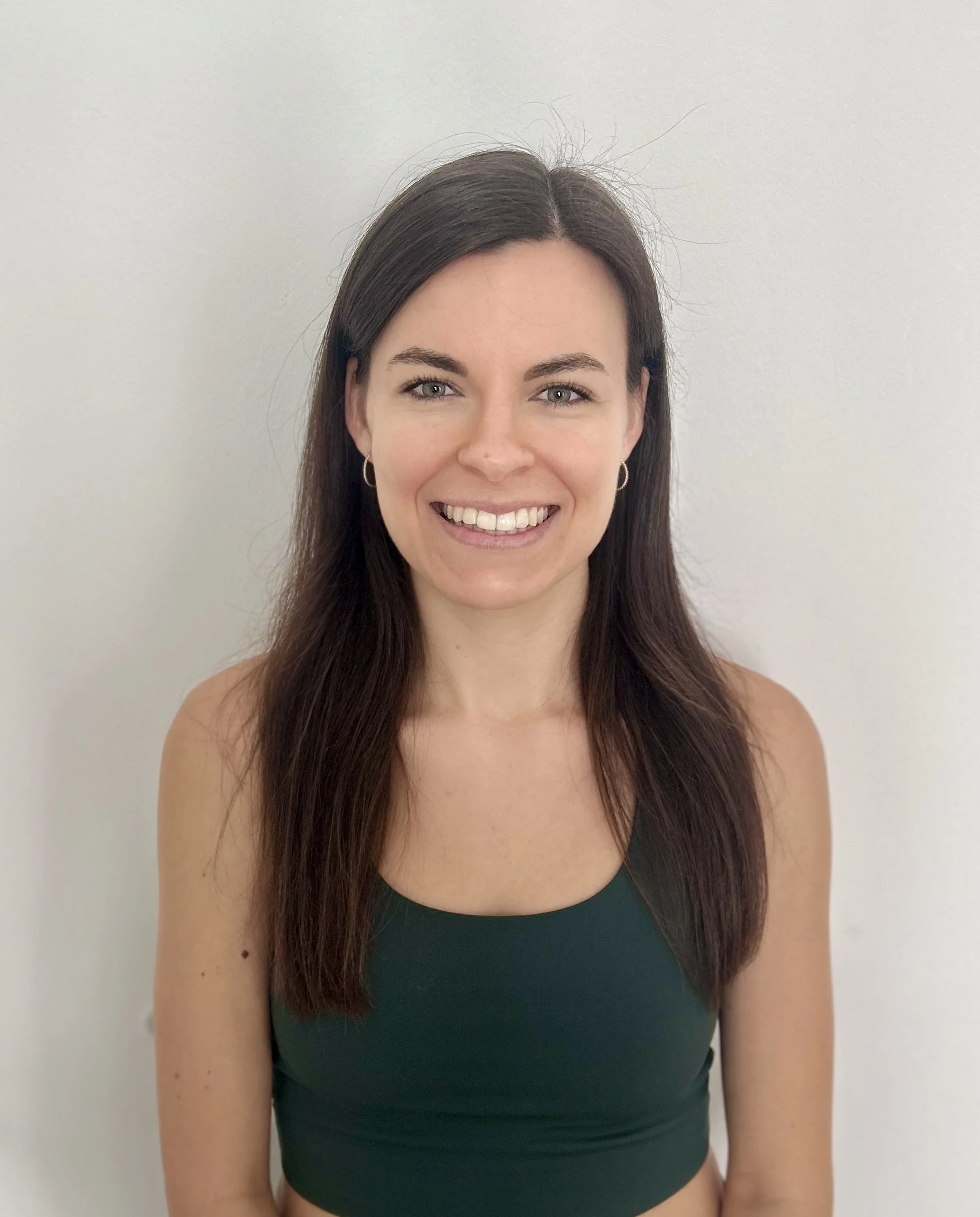 Junge Frau mit langen braunen Haaren, die lächelnd vor einer weißen Wand steht.