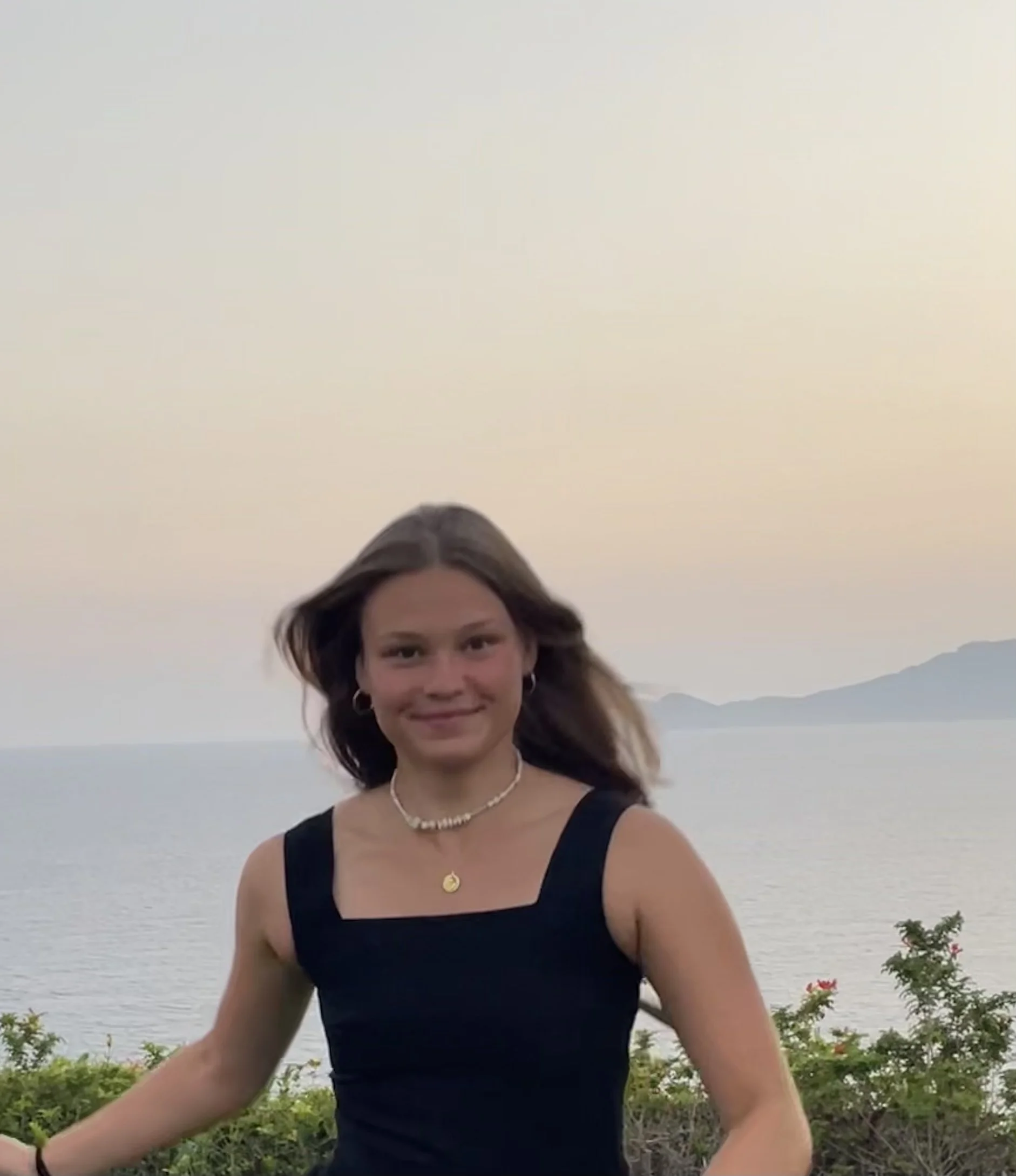 Junge Frau steht vor einer Wasserlandschaft bei Sonnenuntergang, trägt ein schwarzes Kleid, Schmuck und lächelt.